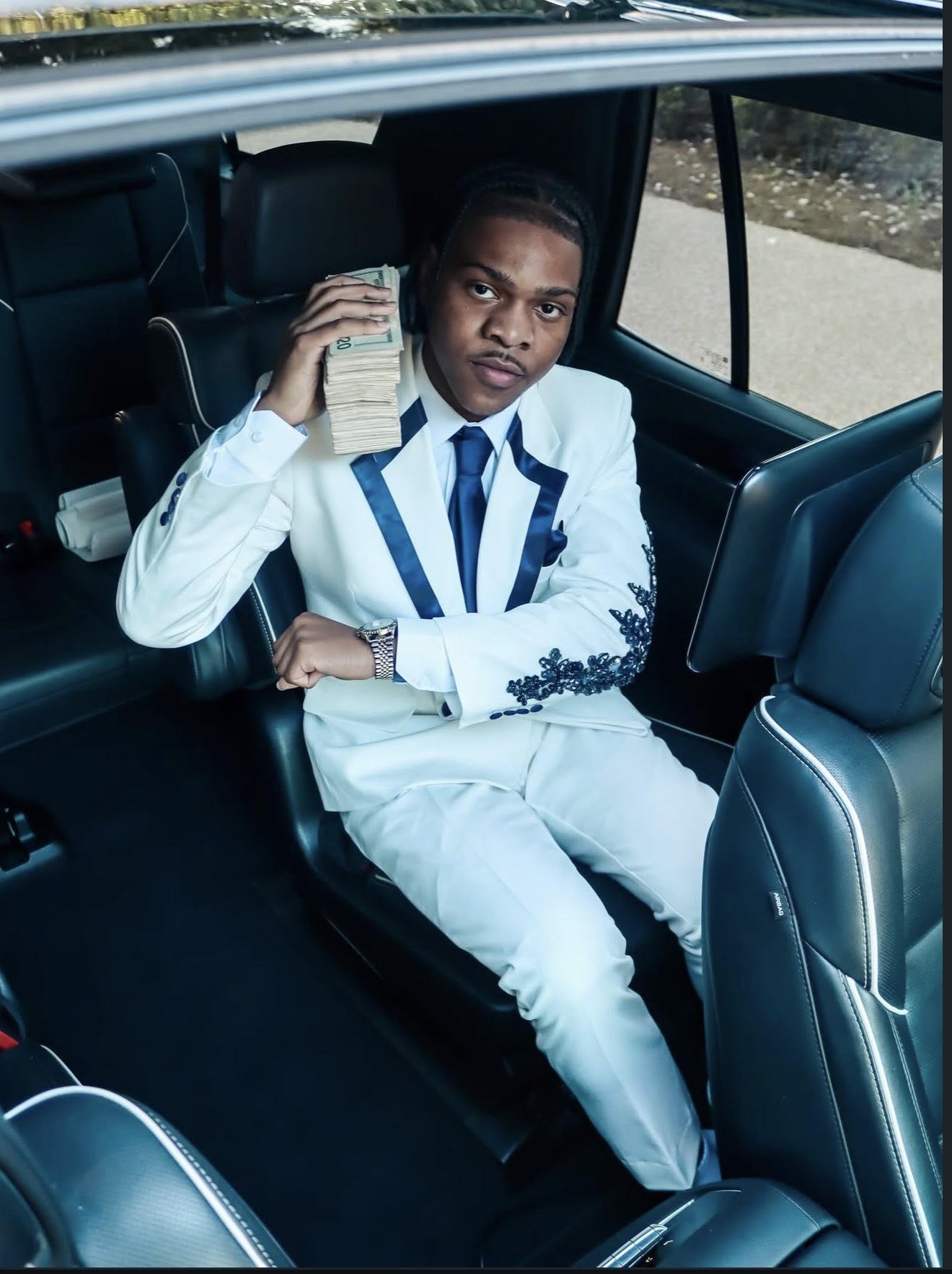A young man in a white suit with navy blue accents sitting in the back seat of a car, holding a stack of U.S. dollar bills to his ear like a phone, with a confident expression.