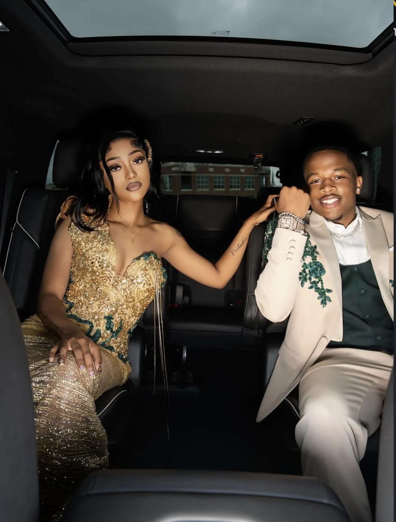 A young woman and man dressed elegantly sitting inside a car, with the woman in a gold dress and the man in a cream suit, smiling and looking at the camera.