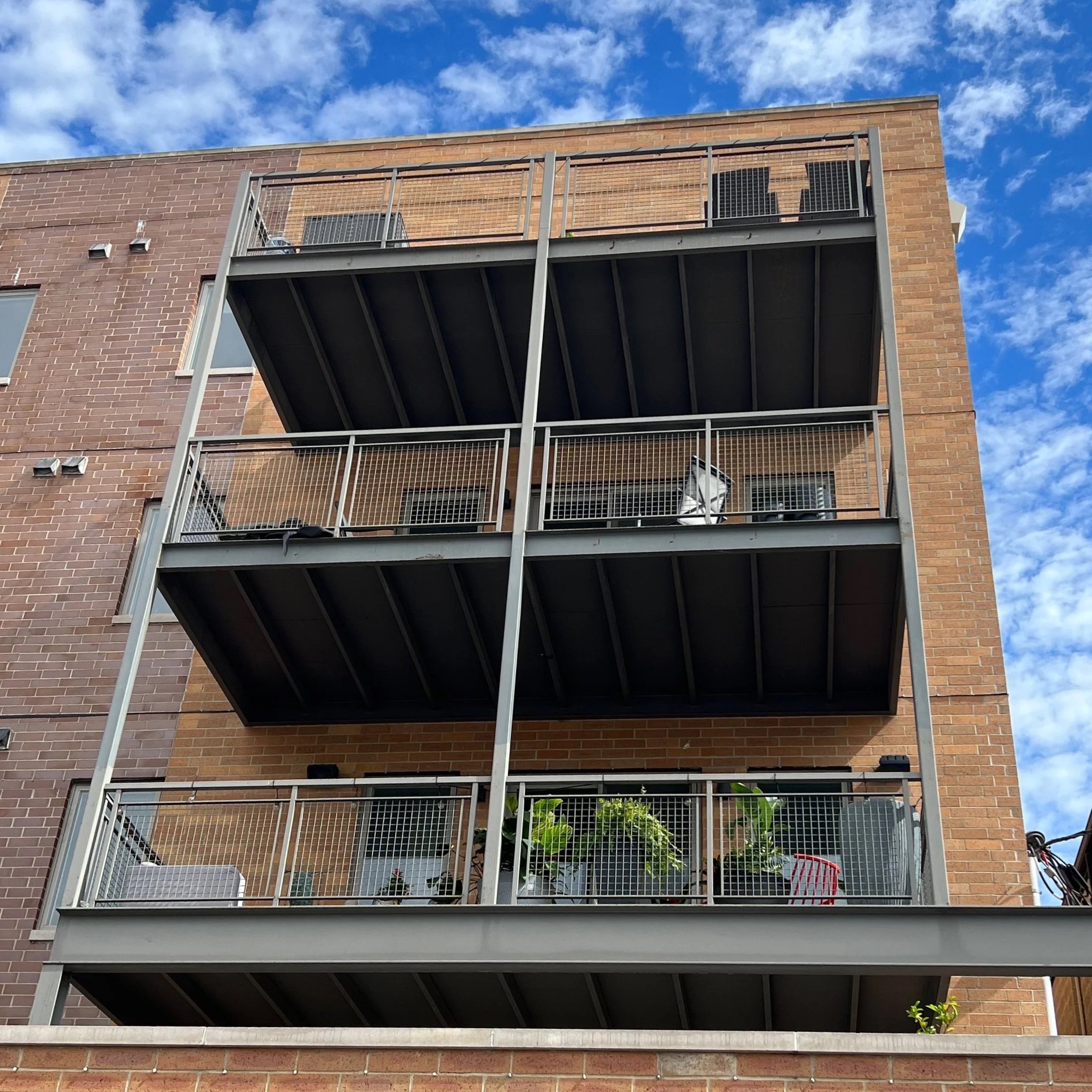 View of a multi-story brick building with metal fire escape balconies, against a blue sky with some clouds.