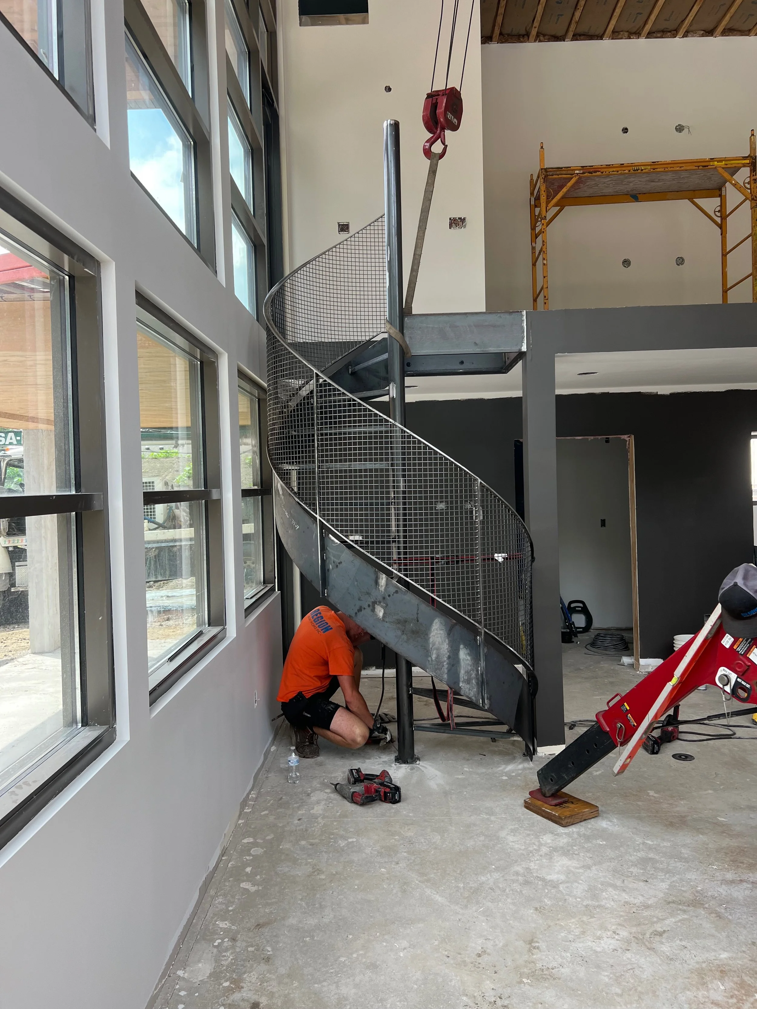 Worker installing a spiral metal staircase inside a building under construction. The staircase is attached to a black column and has a safety cage. Construction tools and equipment are scattered around, including a drill, a water bottle, and a red de