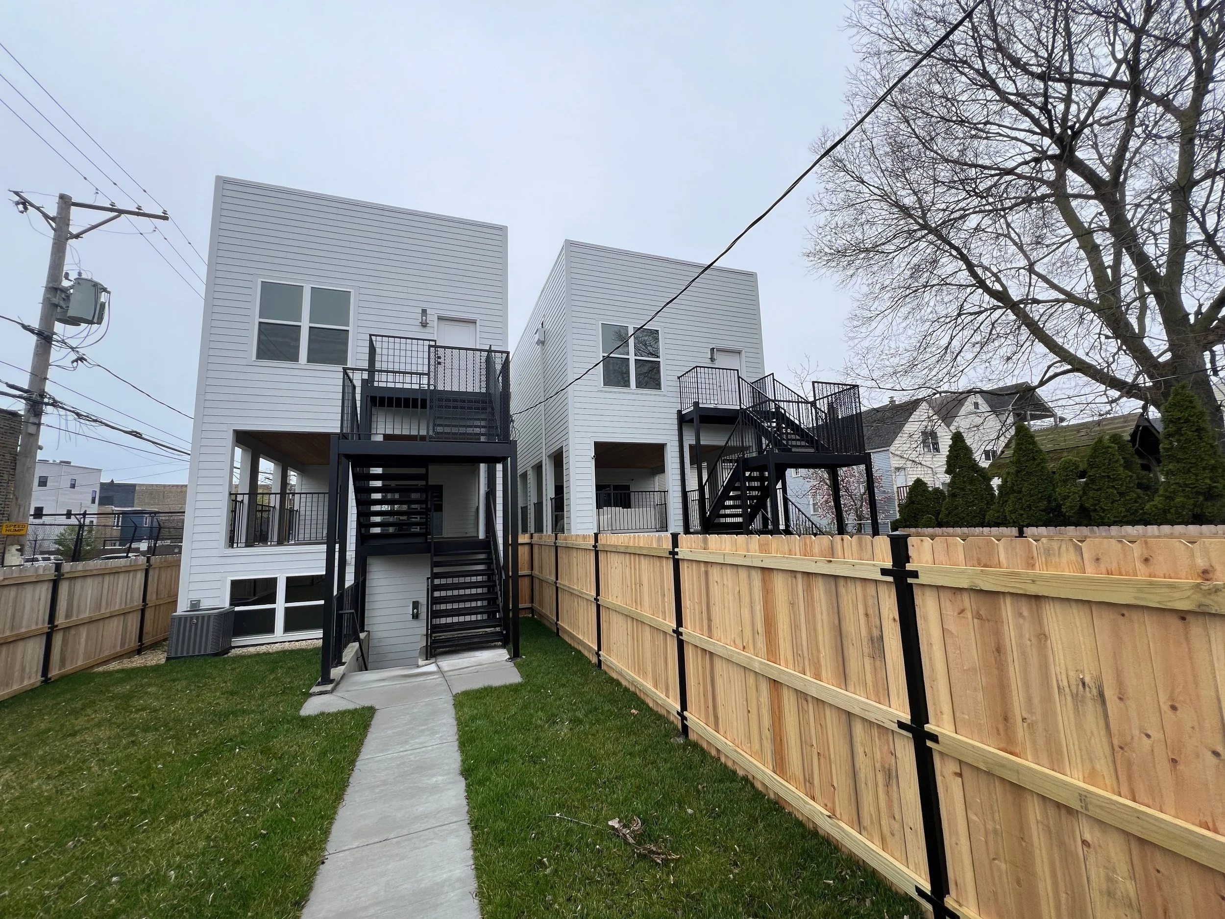 Backyard with grass, paved walkway, wooden fence, a modern white multi-story building with staircases and balconies, electrical pole with wires, and leafless trees.