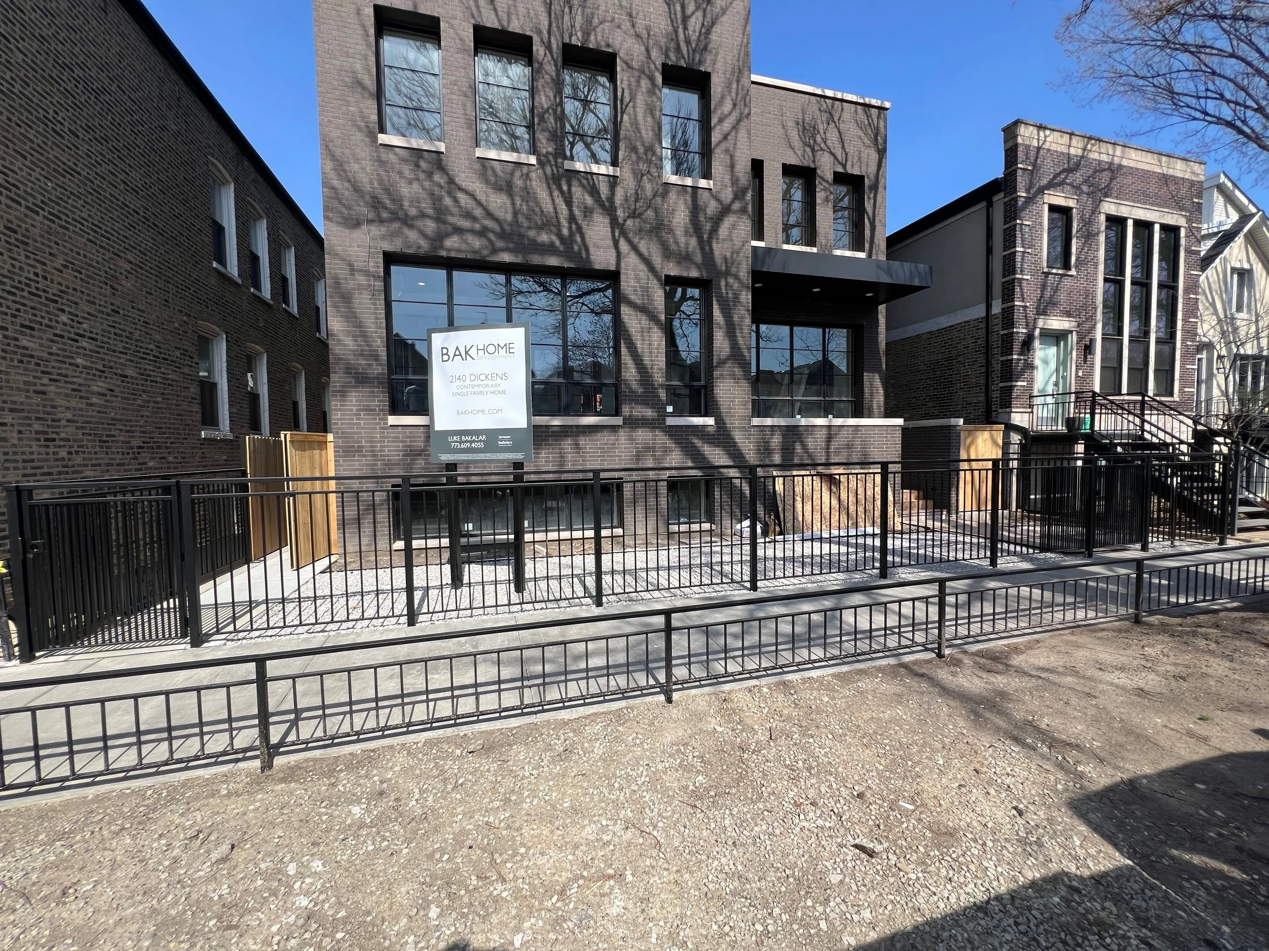 Modern multi-story brick building with large windows and a sign that reads "BAK HOME DEVELOPMENT, 2140 DICKENS, CONTEMPORARY, SINGLE FAMILY HOME" in front of the building. Shadow of trees visible on the building facade. Black metal fence and dirt gro