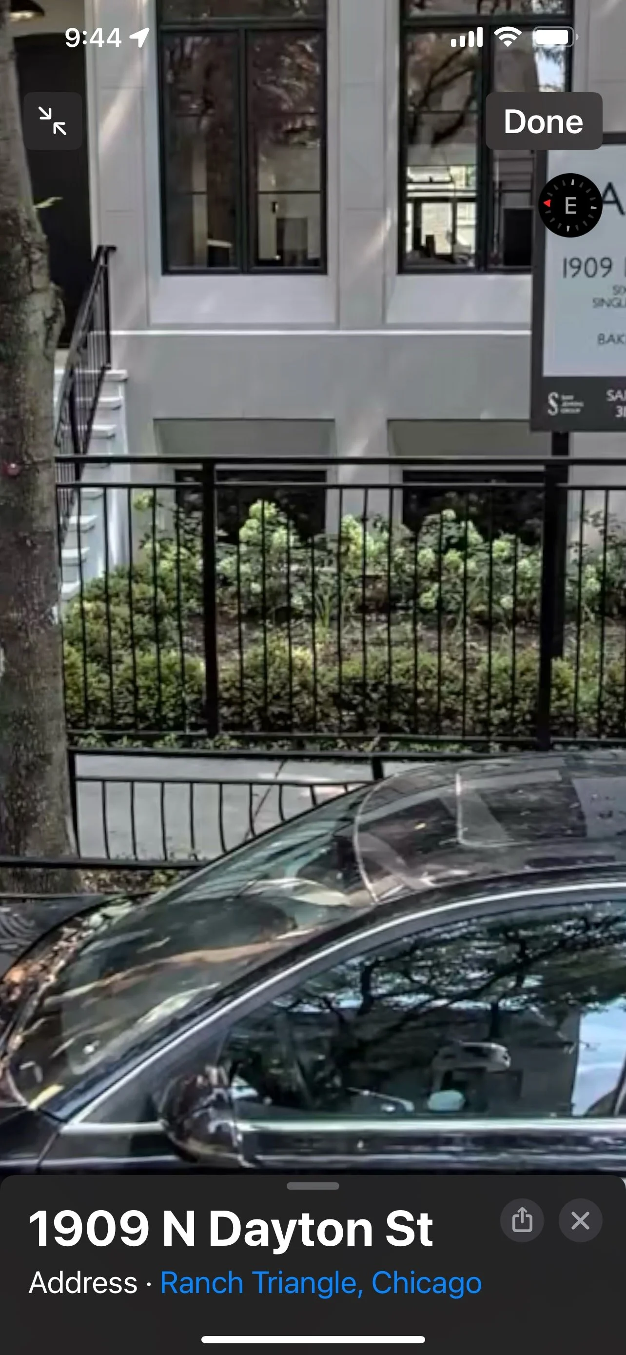 Photo of a street corner showing a building with large windows, a black metal fence, a sidewalk with some plants, and part of a parked car in the foreground. A street sign reads '1909 N Dayton St', located in Ranch Triangle, Chicago.