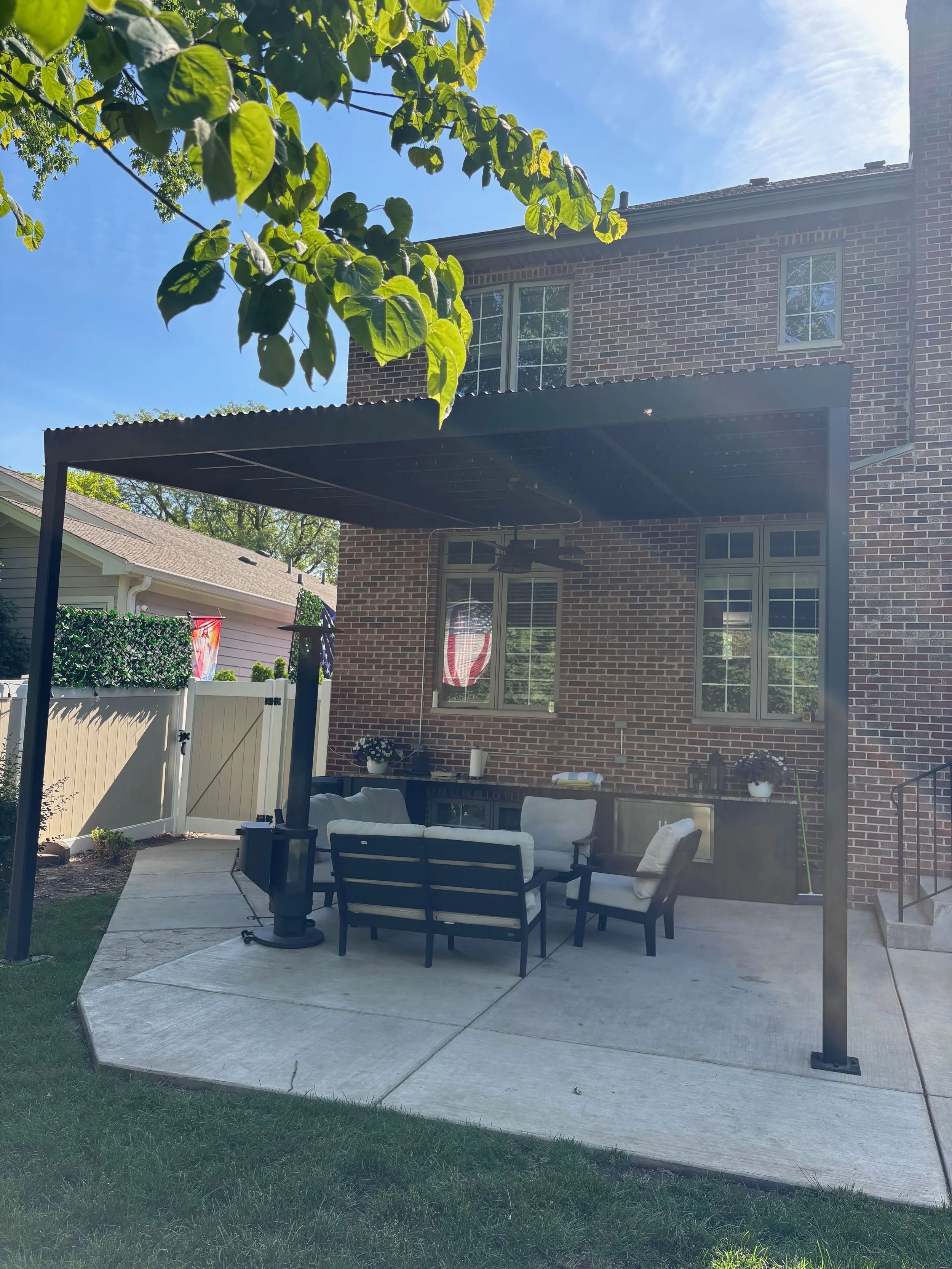 A backyard patio with outdoor furniture includes a sofa and chairs with beige cushions, a black table, and a ceiling fan underneath a black metal pergola. The patio is surrounded by a white fence, and there are trees and a brick house in the backgrou