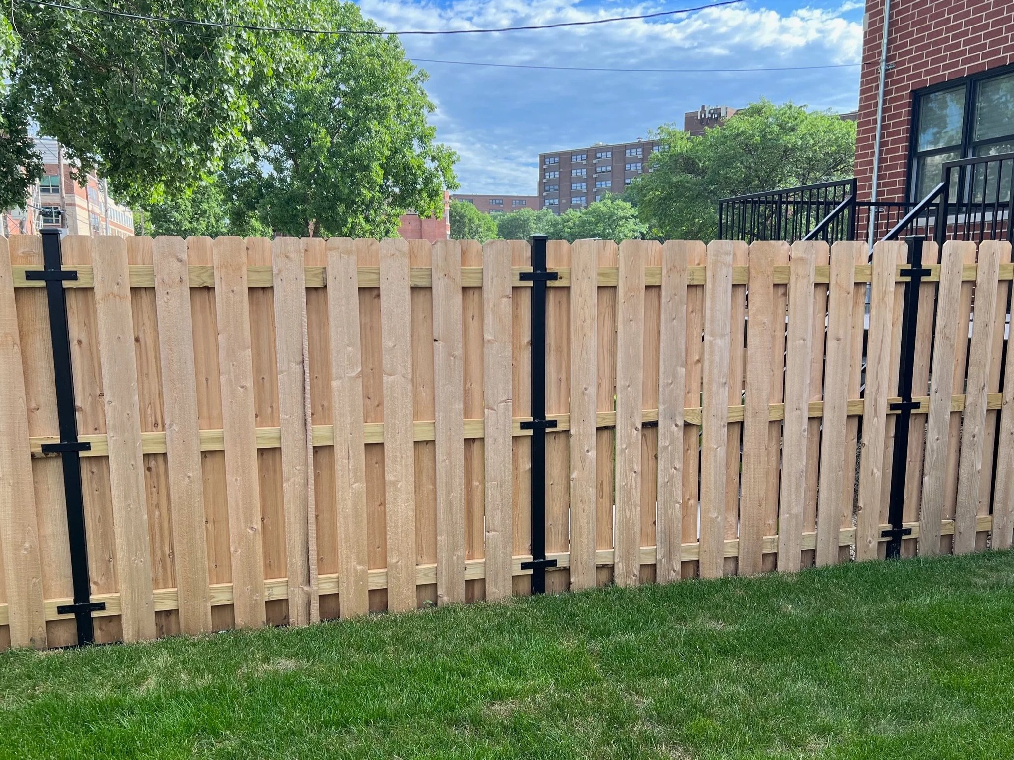 A wooden privacy fence with black metal supports in a backyard, with green grass in the foreground, trees and buildings in the background, and a partly cloudy sky.