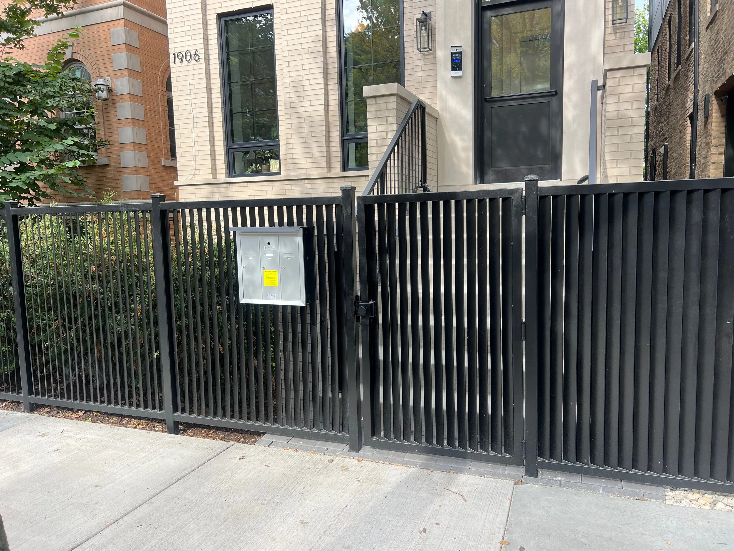 View of a modern residential building entrance with a black metal gate, mailbox, stairs, light fixtures, and large windows, with a tree on the left and neighboring buildings on the right.
