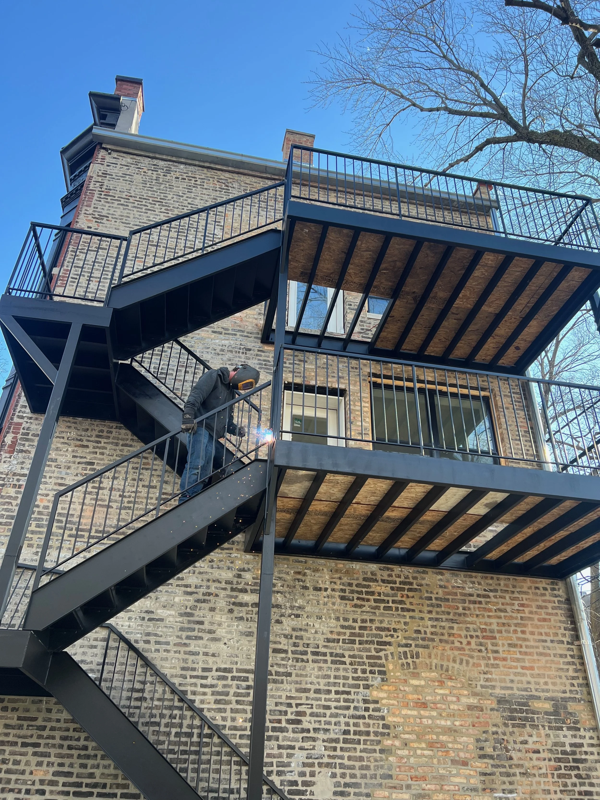A person welding on a black metal staircase outside a brick building. The staircase leads to a second-floor balcony. The sky is clear and blue, and a large, leafless tree is visible in the background.