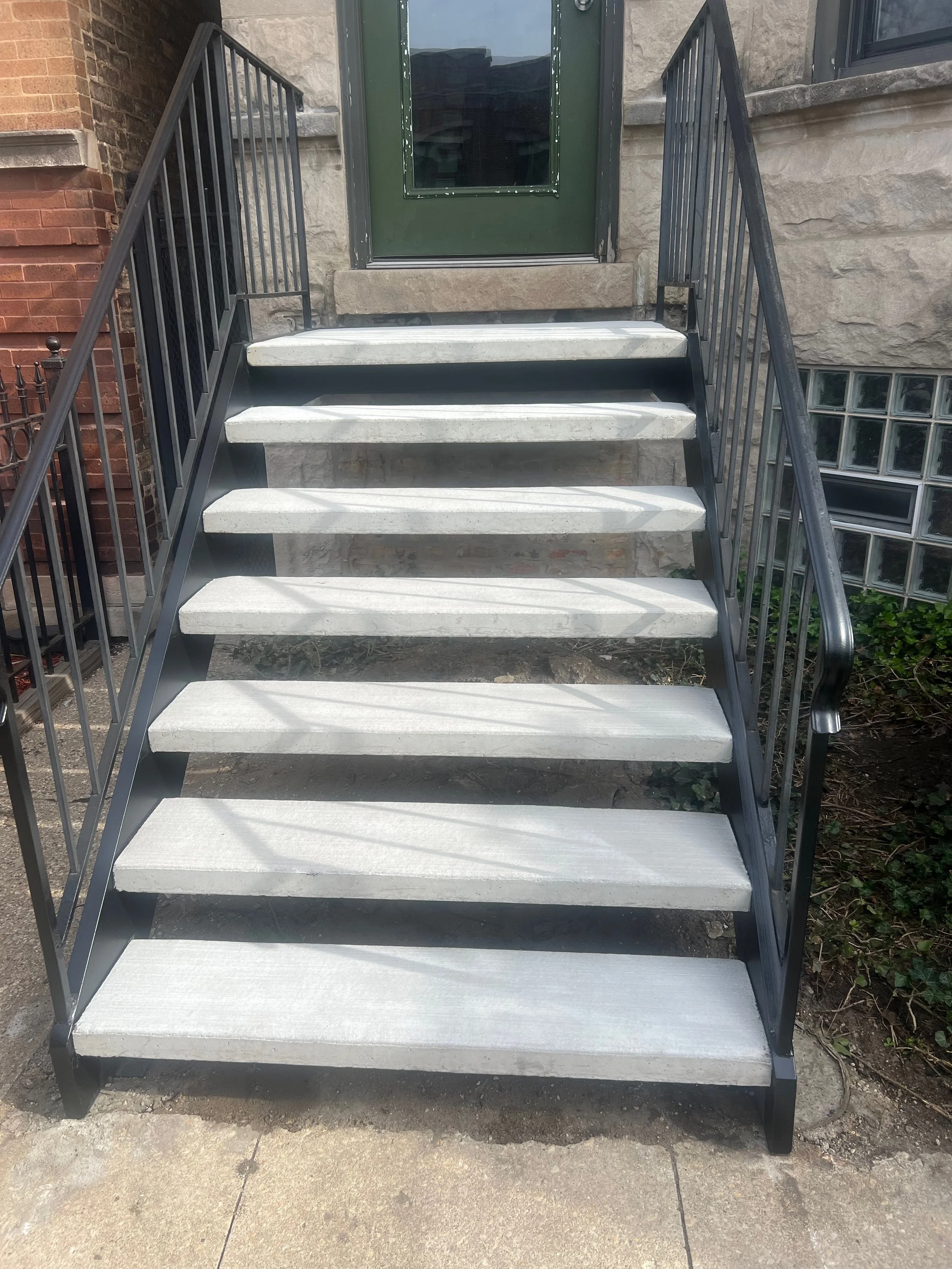 A short outdoor stairway with gray steps, black handrails, leads to a green door with a glass window, set into a beige brick and stone building.