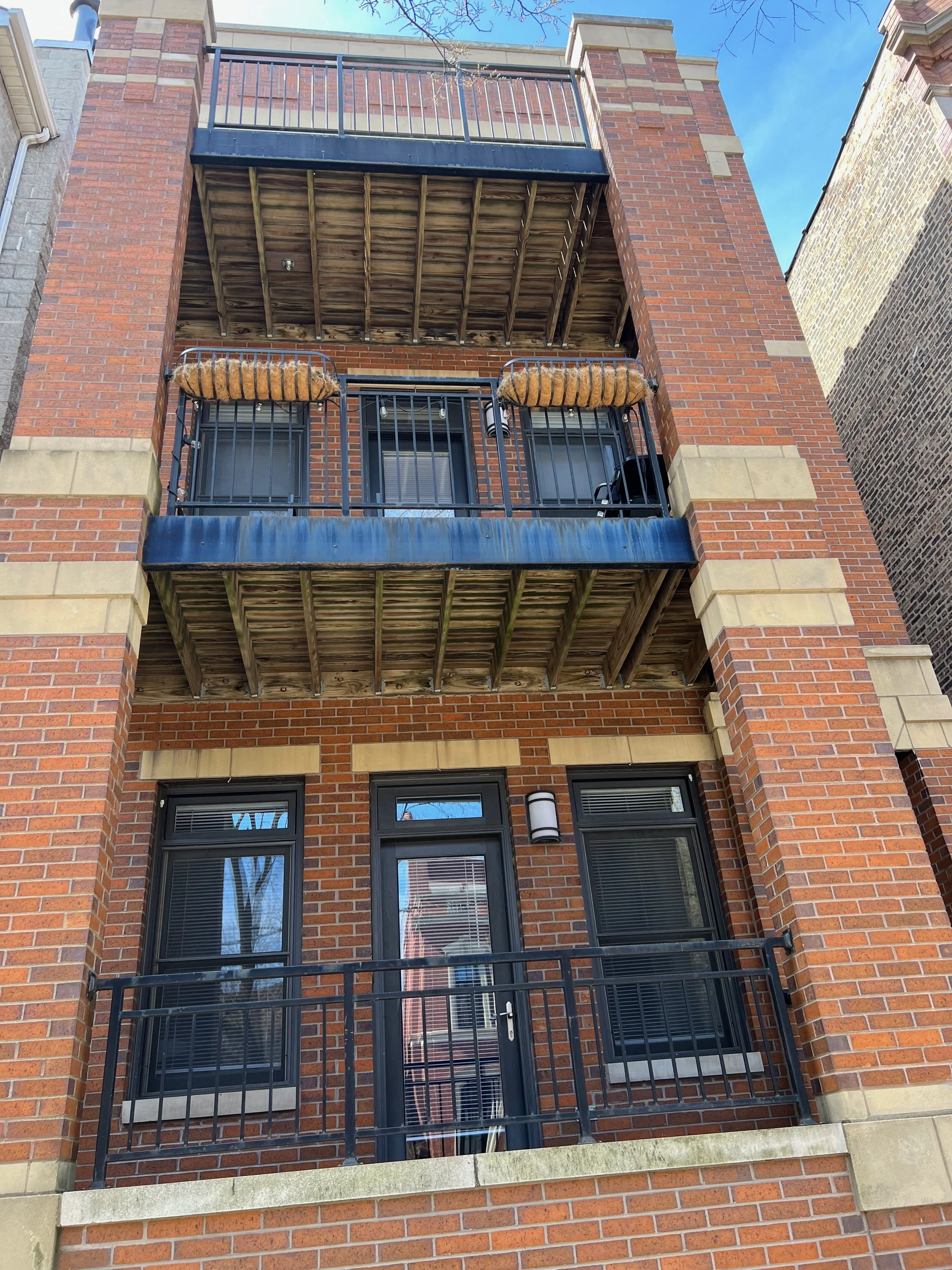 Several balconies in a brick apartment building with black metal railings and wooden flooring, with some pillows on two balconies, blue sky, and neighboring buildings visible.