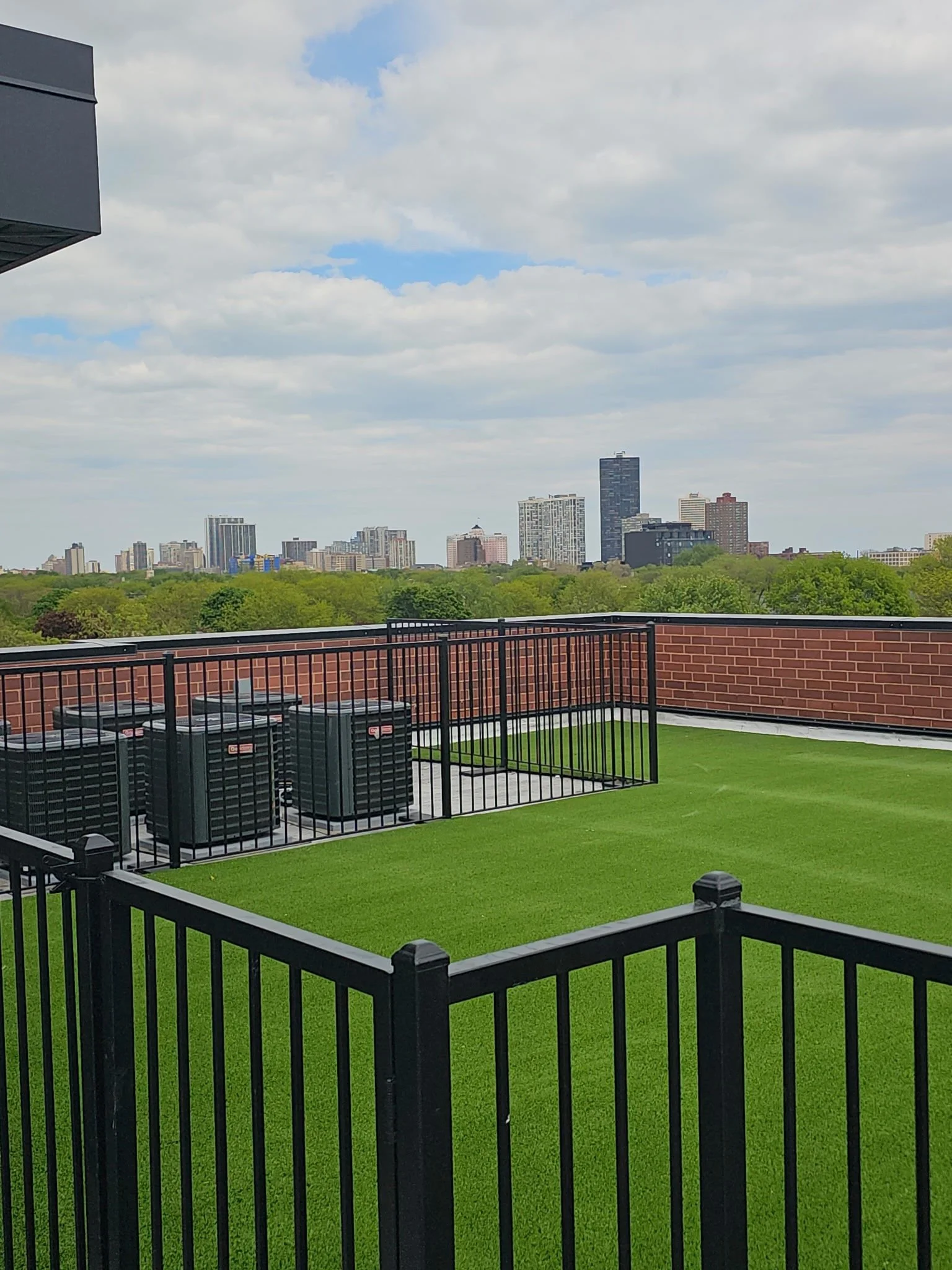 View from a balcony showing green artificial turf, a black metal railing, and several HVAC units. Beyond, a city skyline with high-rise buildings and a partly cloudy sky.