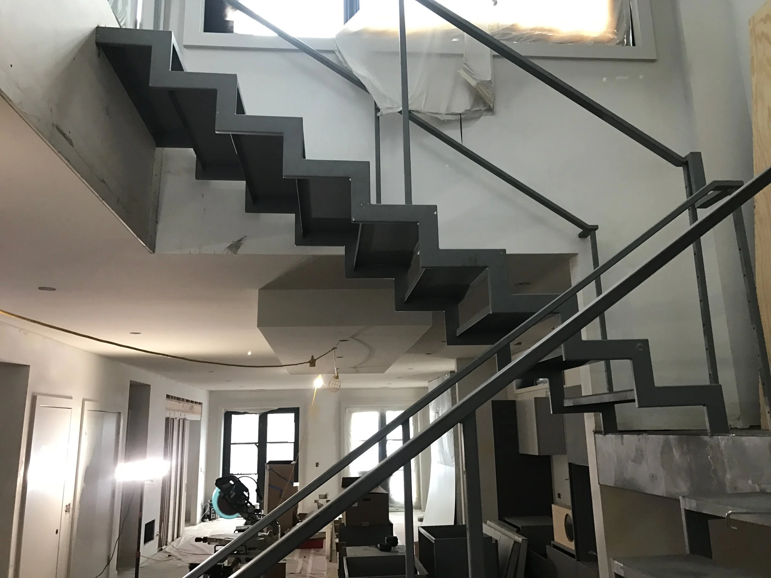 Interior view of a house under construction or renovation, featuring black staircases with exposed metal railings, a vacuum or blower in the foreground, and construction materials and tools scattered around. Windows provide natural light.