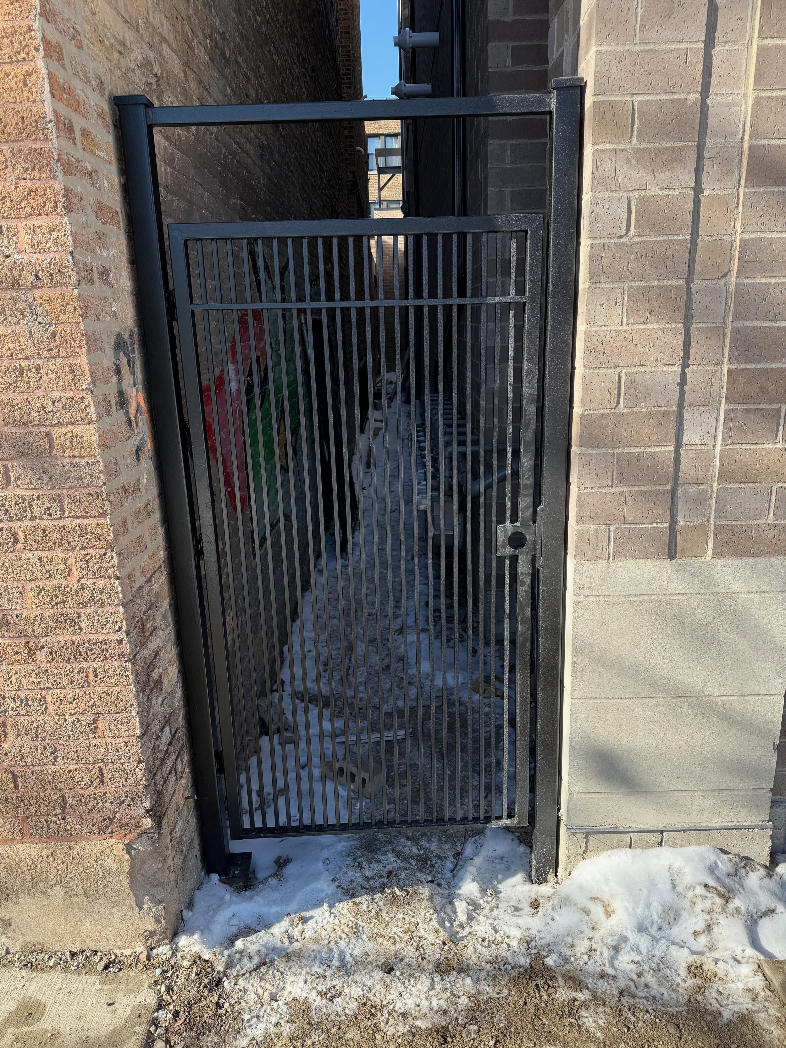 Black metal gate in front of a brick wall with snow on the ground.