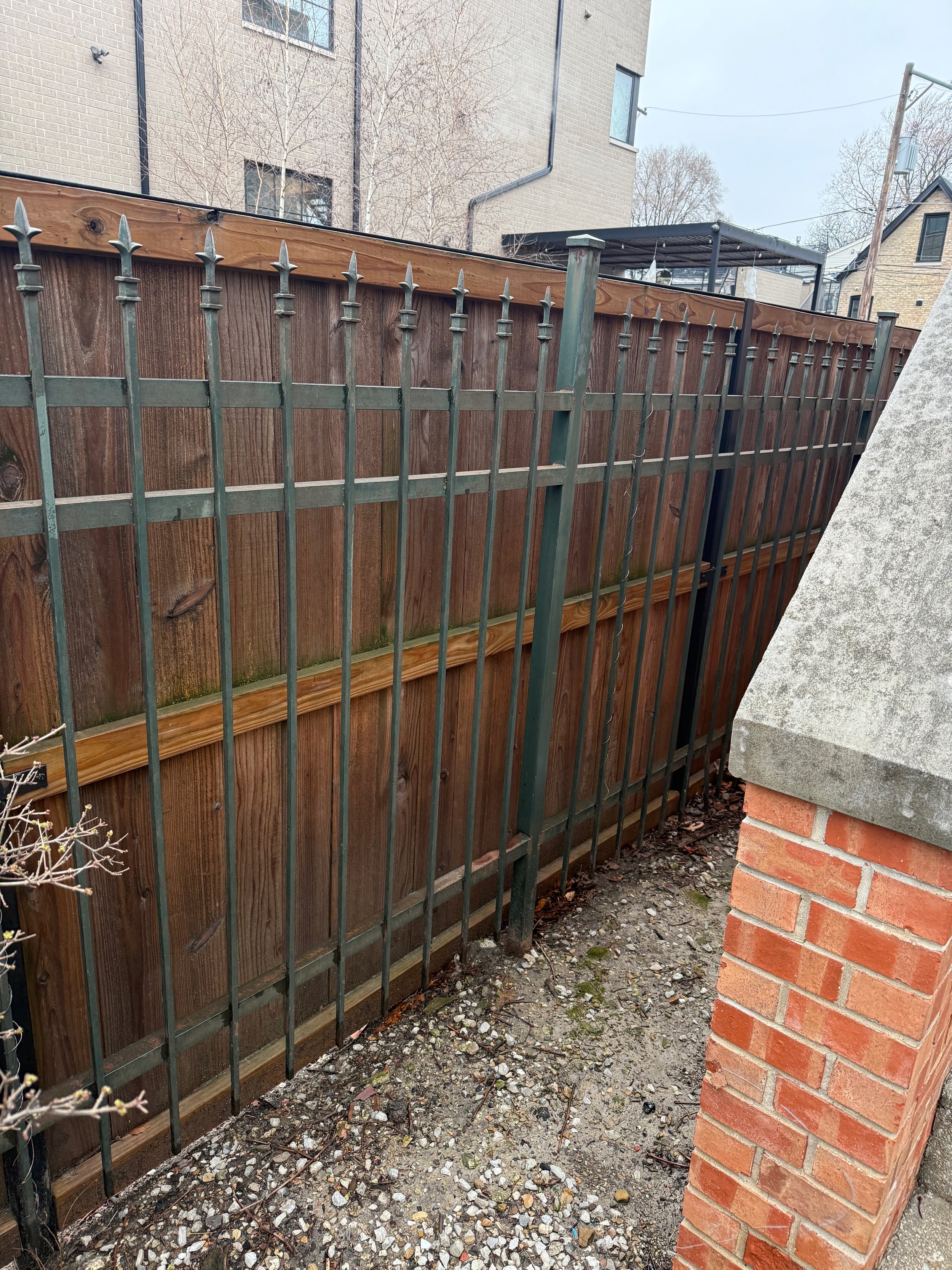 A backyard area with a wooden fence and a metal gate. A brick wall is visible on the right side, and the ground is covered with dirt and small rocks. A pergola structure is in the background, along with nearby houses and a utility pole.