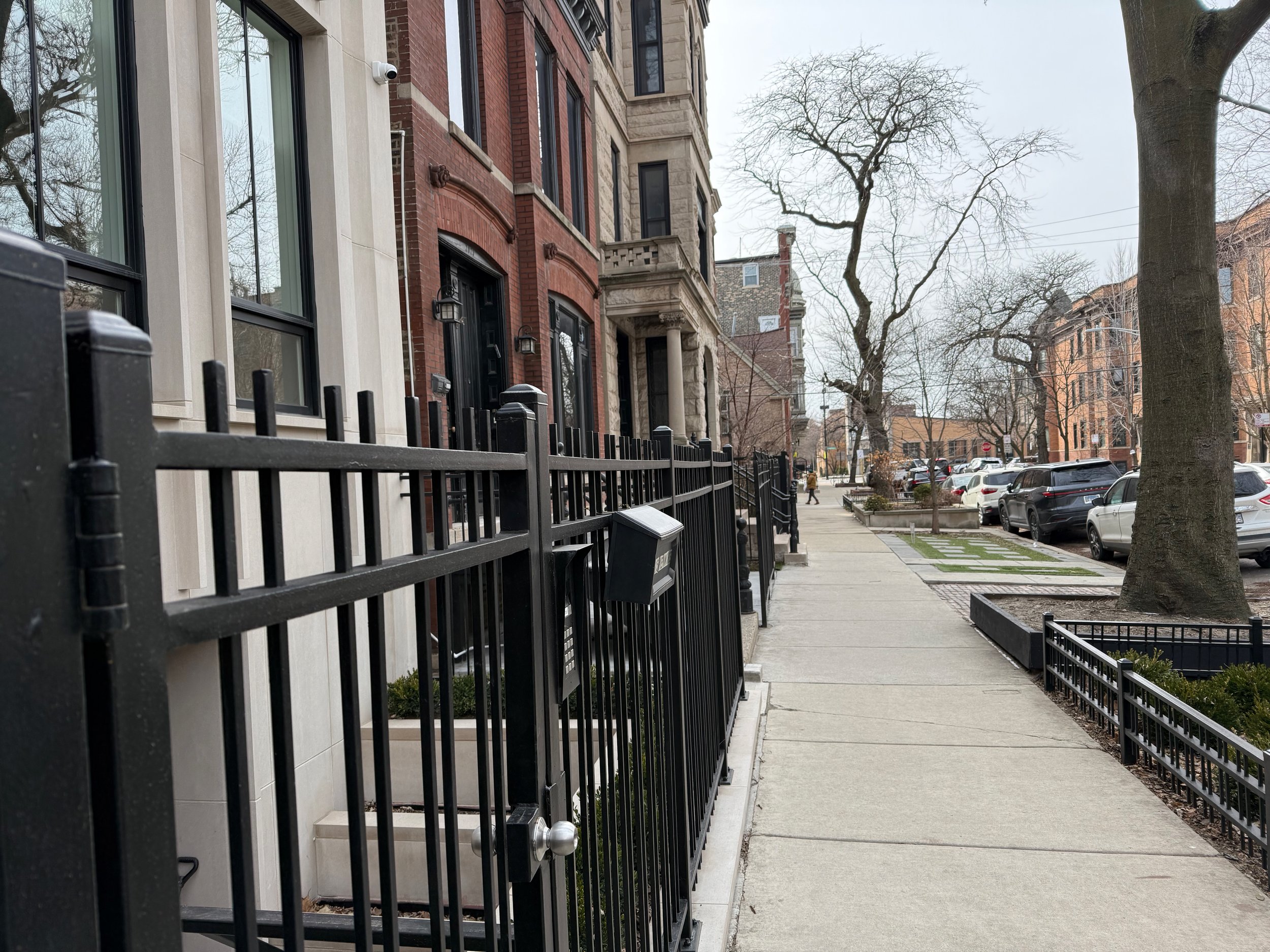 A sidewalk with a black fence and a row of historic-style brick and stone buildings. Bare trees line the street, and cars are parked along the curb.