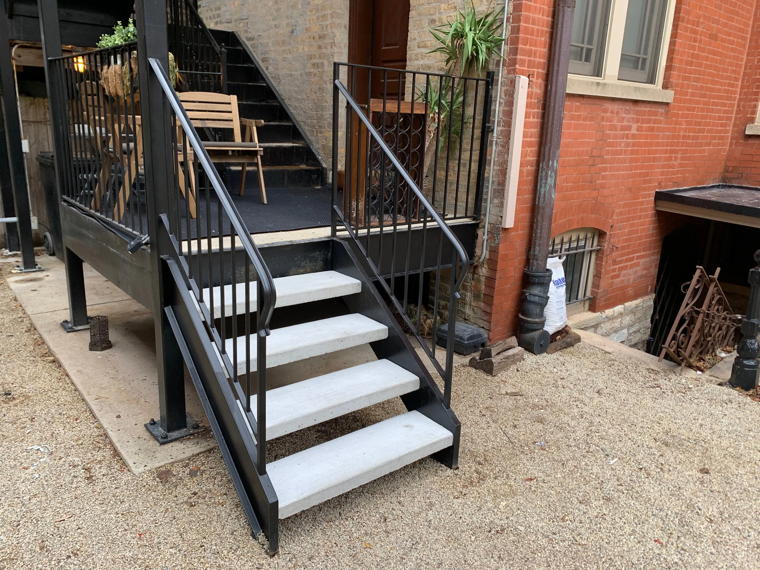 Exterior of a brick building with a metal staircase leading up to a door. Wooden chairs are stacked near the stairs. Potted plant and a discarded bag are nearby.