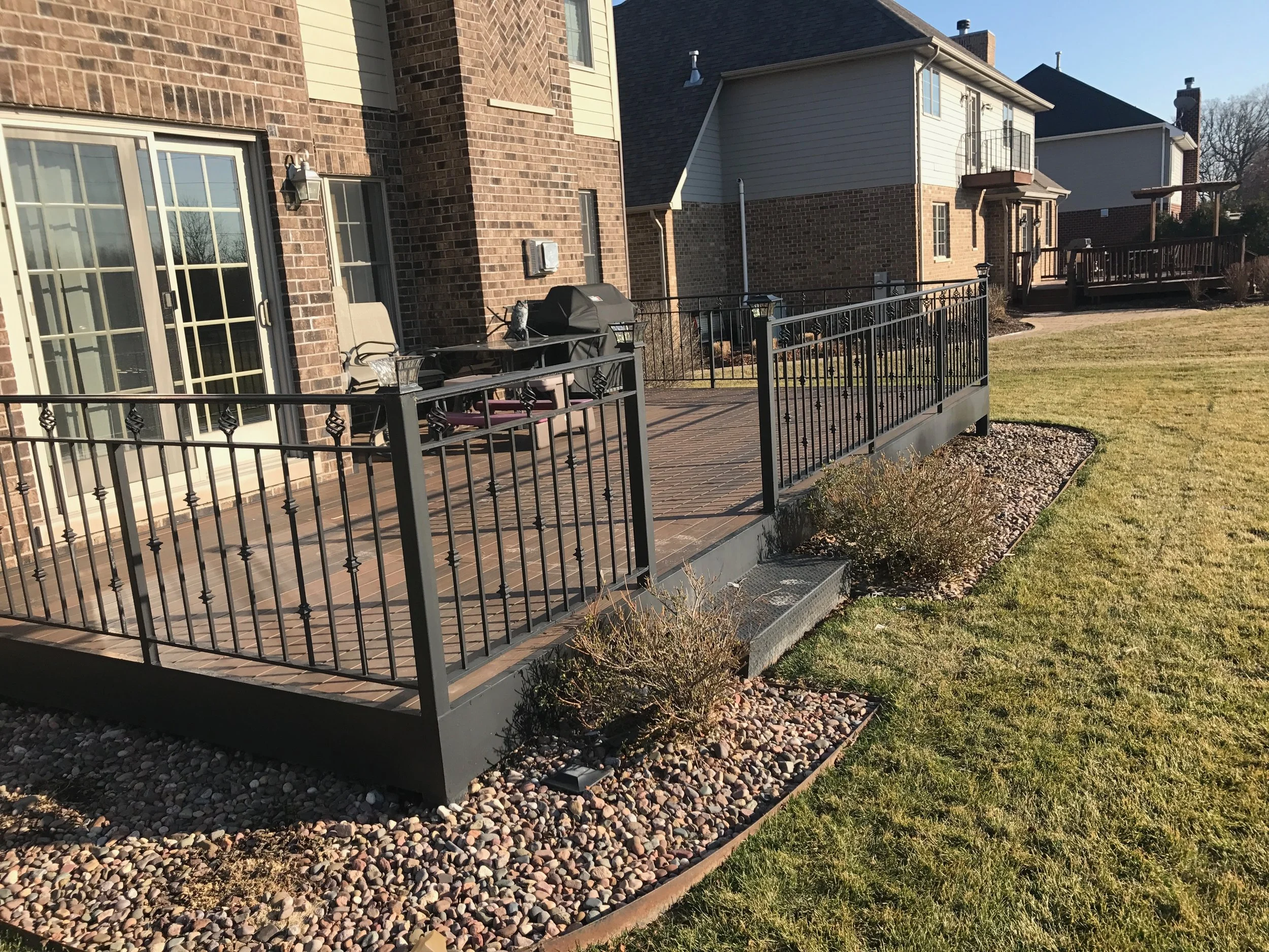 Residential backyard deck with black metal railing, outdoor furniture, barbecue grill, and surrounding landscaping including grass and decorative stones.