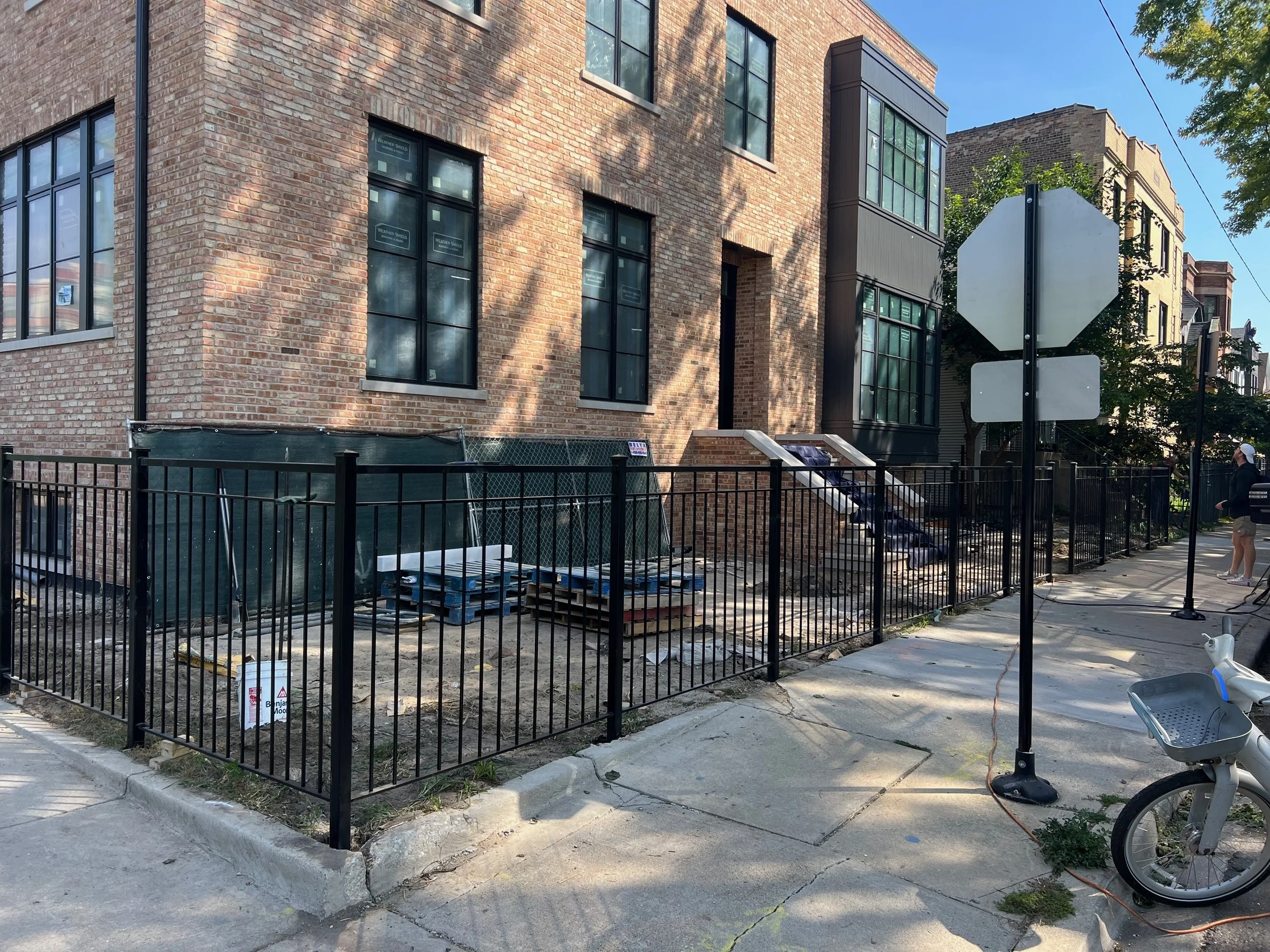 Construction site in front of a brick building with black fencing, construction materials, and a person walking nearby. Traffic signs are visible nearby.