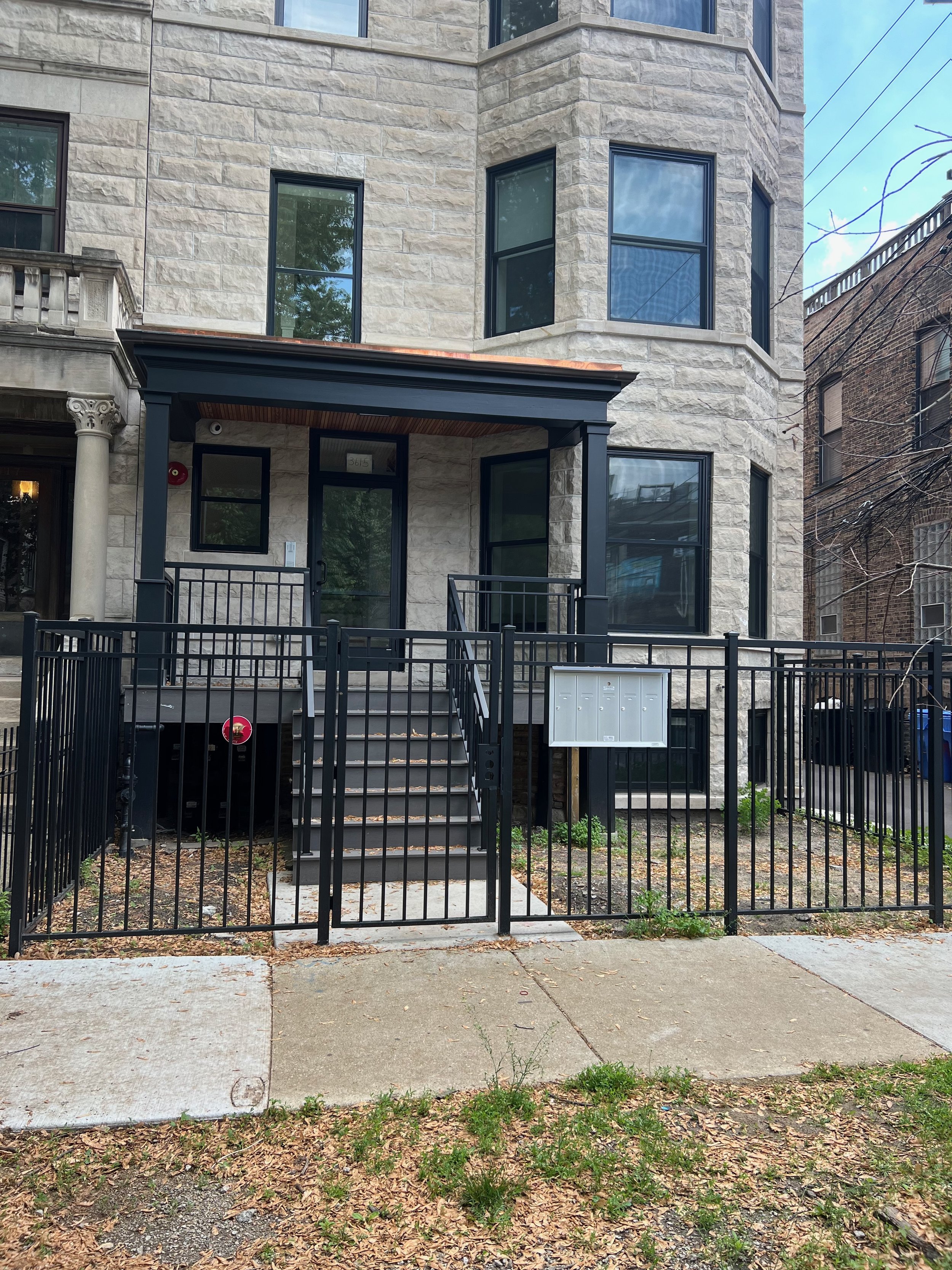 Front view of a modern multi-story apartment building with stone exterior walls, black-framed windows, and a small covered front porch with a black metal railing. There is a black fence surrounding the small front yard and a utility box near the side