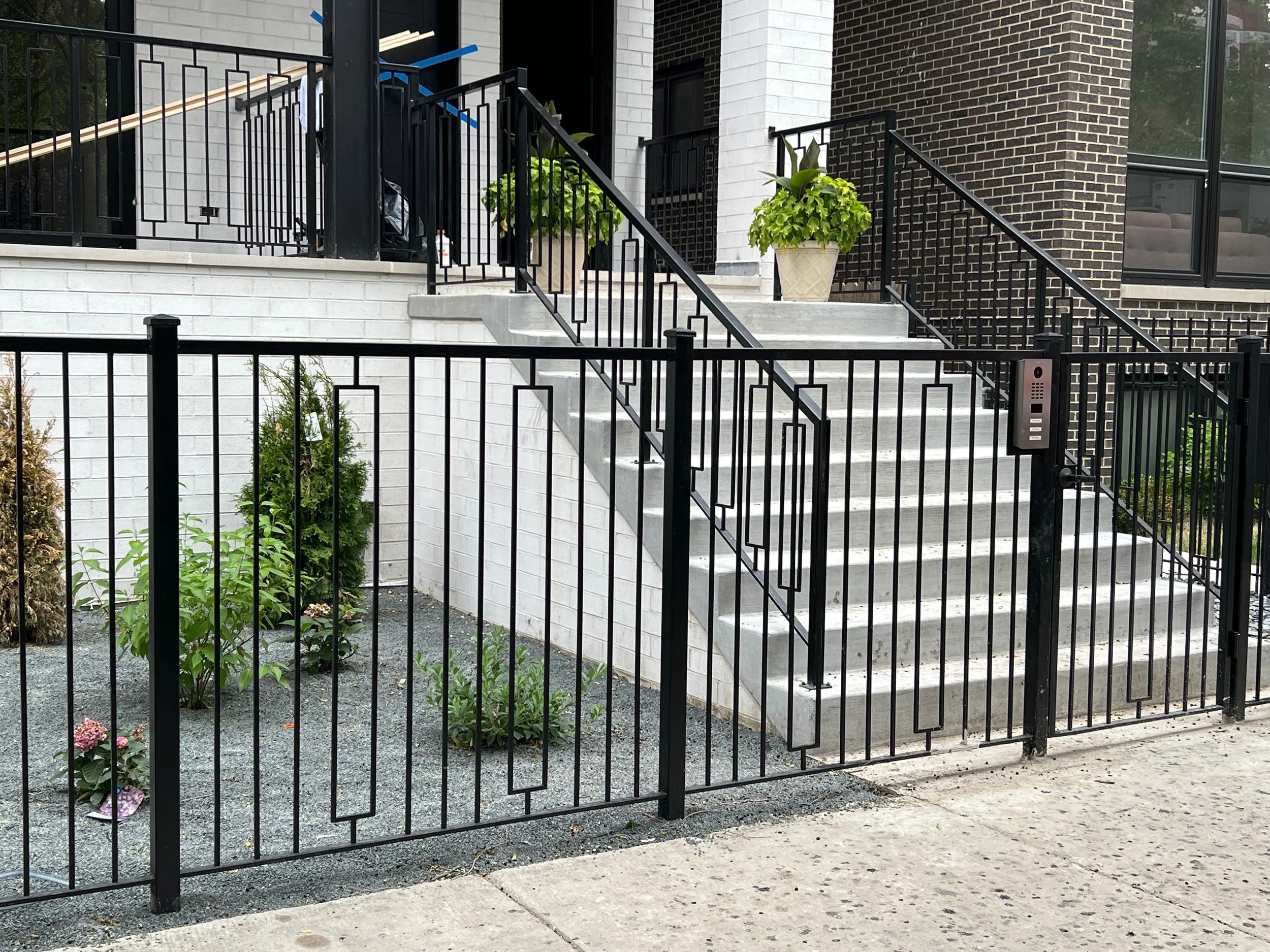 Front view of a modern residential building entrance with concrete stairs, black metal railings, two potted plants, and landscaped garden area.
