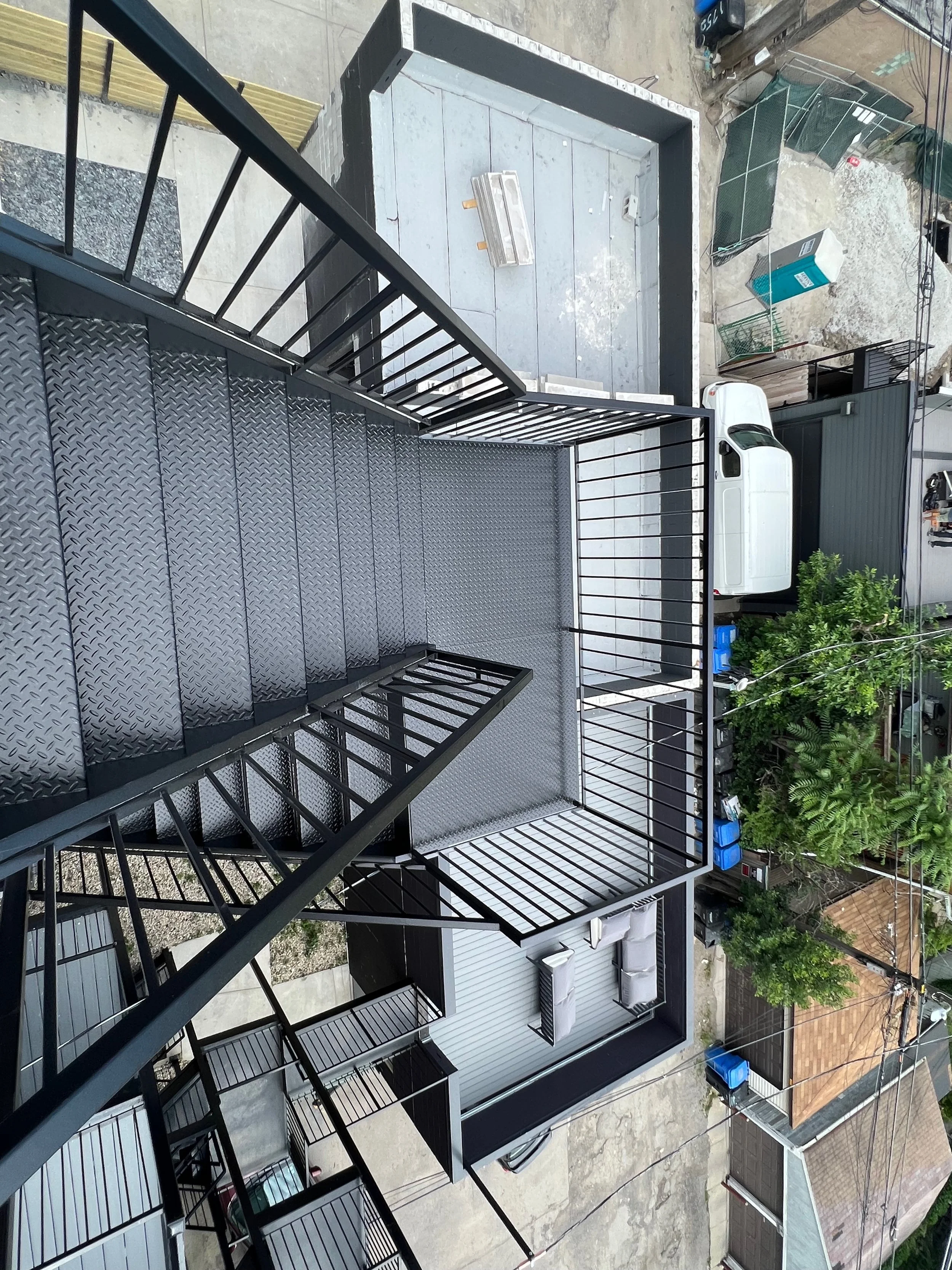 View from a balcony with black metal railings overlooking a parking lot and neighboring buildings, including a white van, some trees, and various outdoor items.