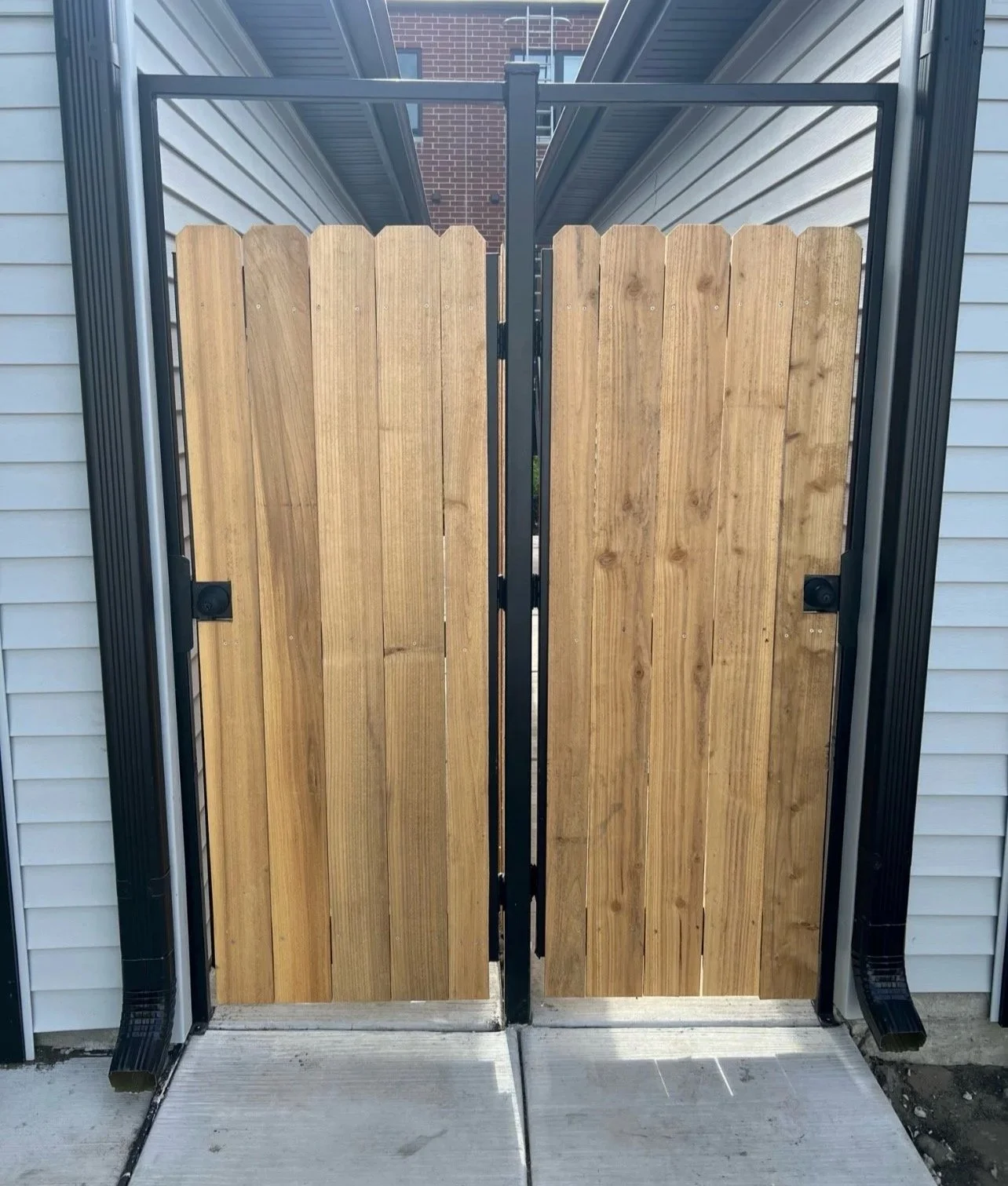 Pair of wooden privacy gates with black metal frames, installed at a residential entrance, flanked by white siding walls.
