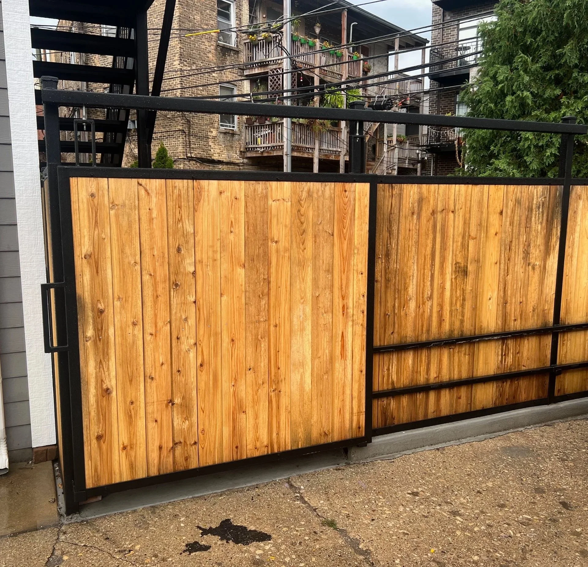 View of a new wooden fence with black metal frame in front of residential brick buildings, some with balconies and hanging plants, and green trees on the right side.