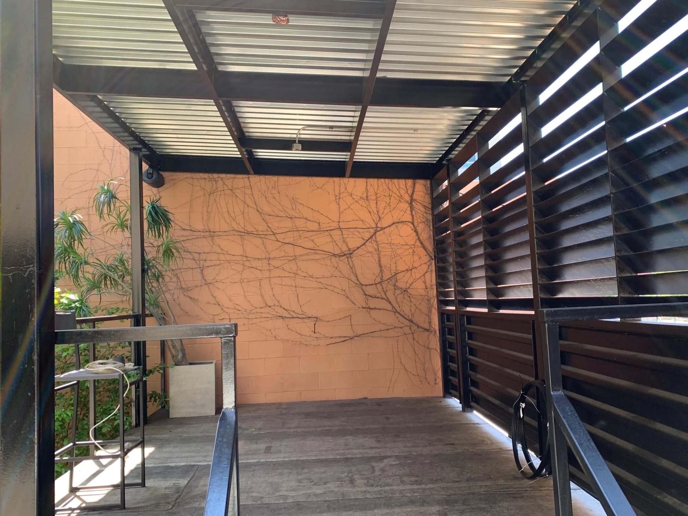 View from below of a covered outdoor patio or balcony with a metal roof, wooden flooring, black metal railings, and a decorative peach-colored wall with vine-like lines. There are some plants and a small table or stand on the left side.