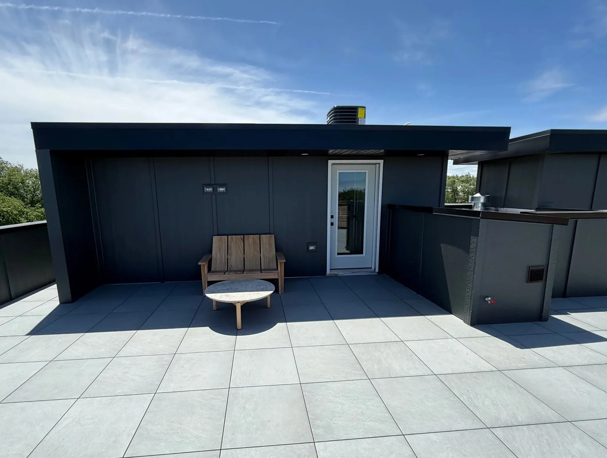 Rooftop terrace with gray tiled flooring, a wooden bench, a small round table, a door, and black railings, under a partly cloudy sky.
