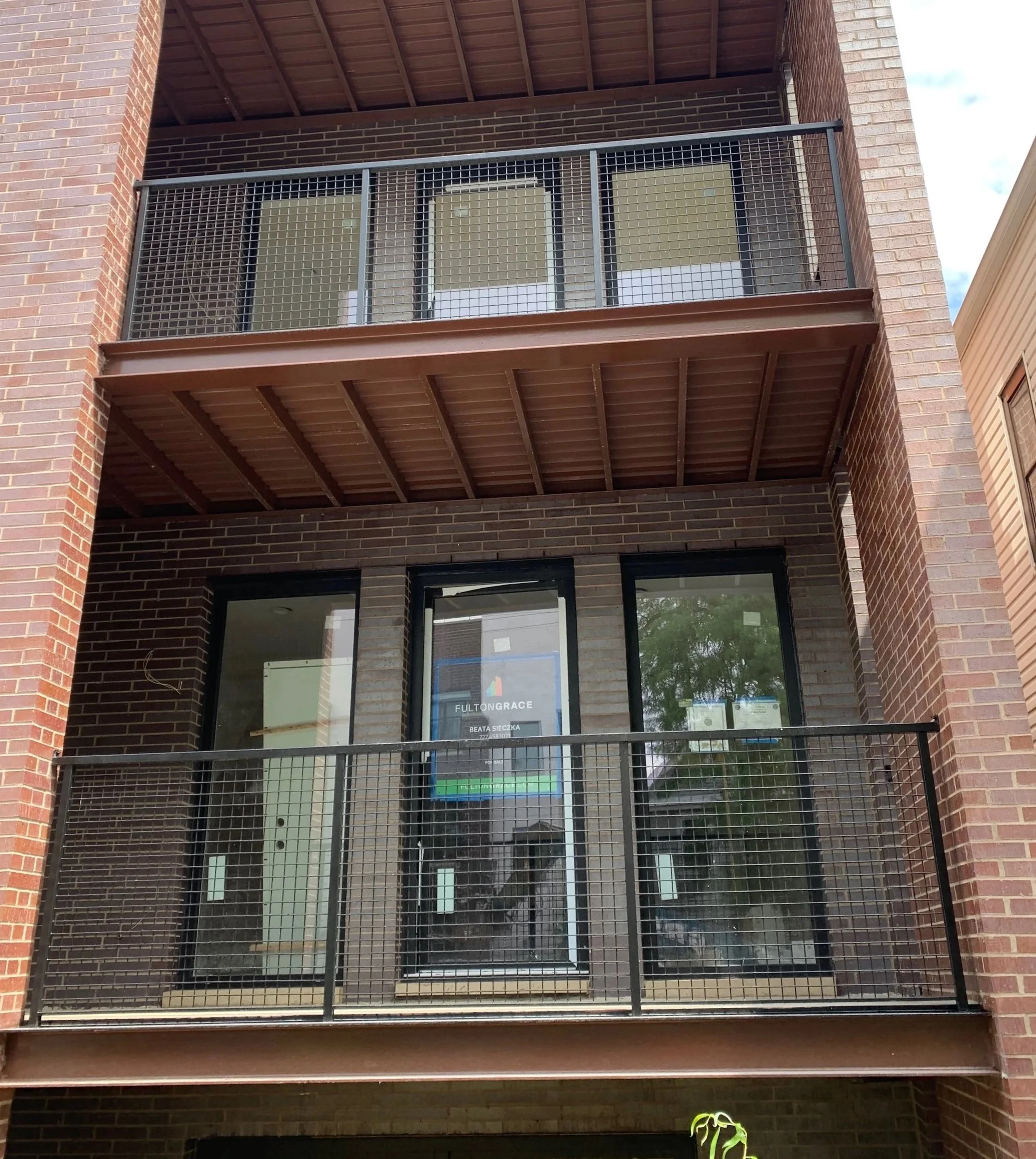 The image shows a modern apartment building door with a sign that reads 'Fulton Grace' and an address, located on a brick wall. The building features two balconies with metal railings, and the lower balcony has a pair of bright green shoes at the bot