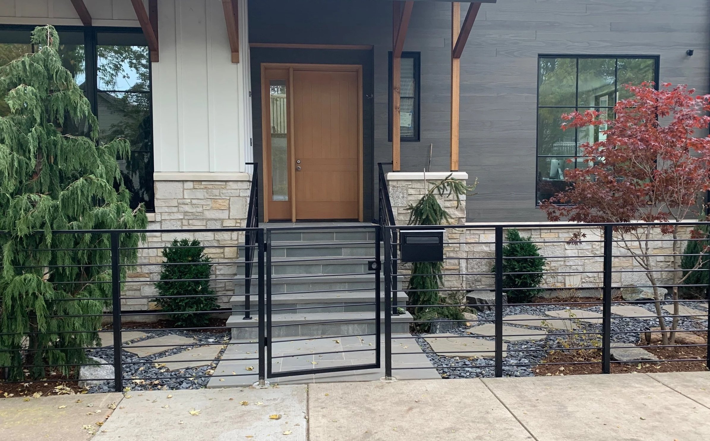 Front view of a modern house with steps leading up to a wooden front door, large windows, two small bushes, a red maple tree, and green foliage on the left side, surrounded by a black metal fence.
