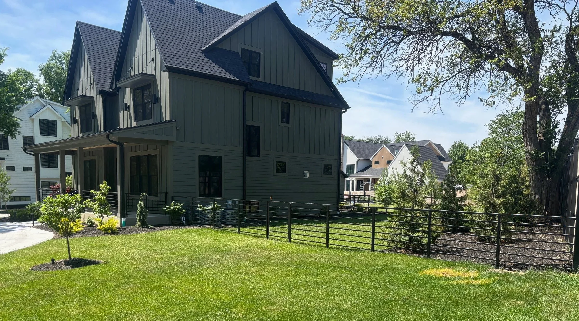A modern, multi-story house with vertical siding in a residential neighborhood, surrounded by a well-maintained grassy yard with young trees and a black fence, under a clear blue sky with a large tree in the foreground.