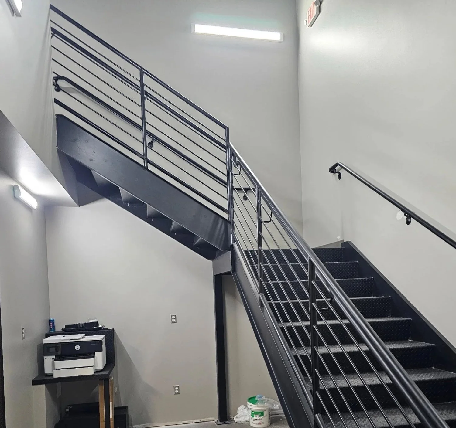 Interior stairwell with metal staircase and handrails, white walls, some office equipment, a small table with a printer, a bucket, and cleaning supplies.