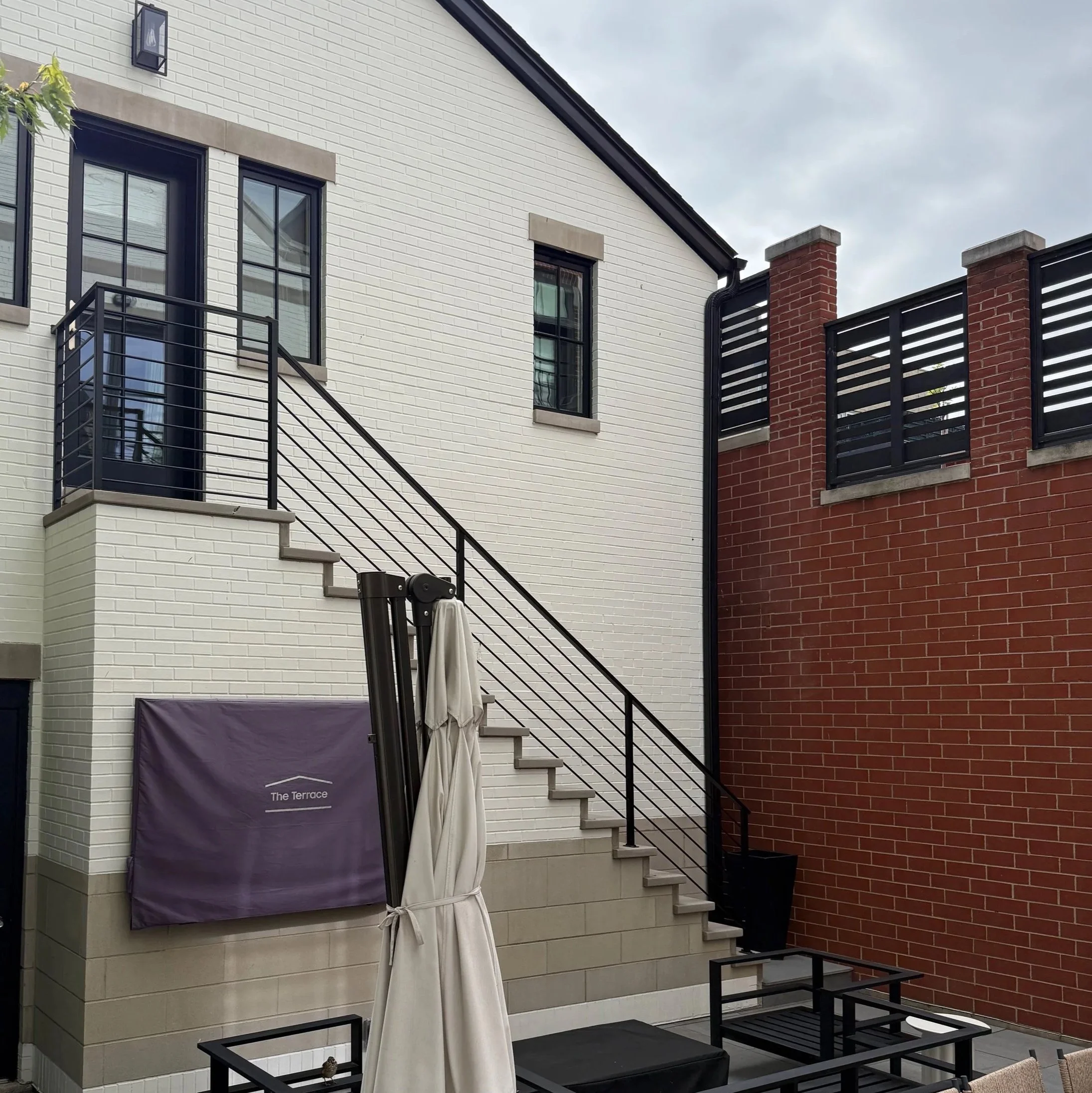 Exterior view of a modern residential building with white brick walls and a red brick wall, black window frames, and metal railings on a small outdoor balcony area under an overcast sky.