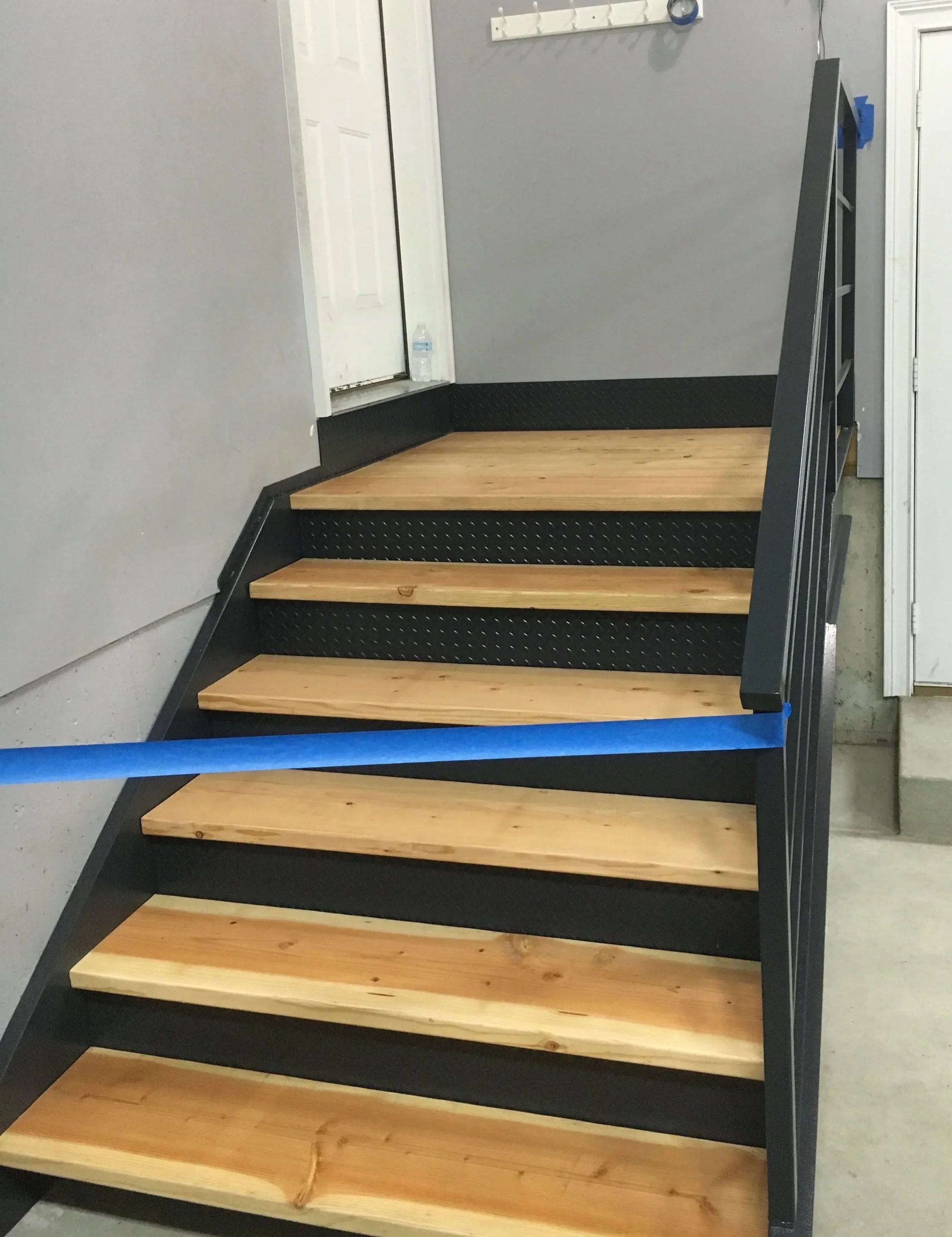 A staircase with four wooden steps, a black metal frame, black diamond-patterned metal risers, and a black handrail on the right side. The staircase is against a gray wall with a white door and a water bottle on the floor next to the door.