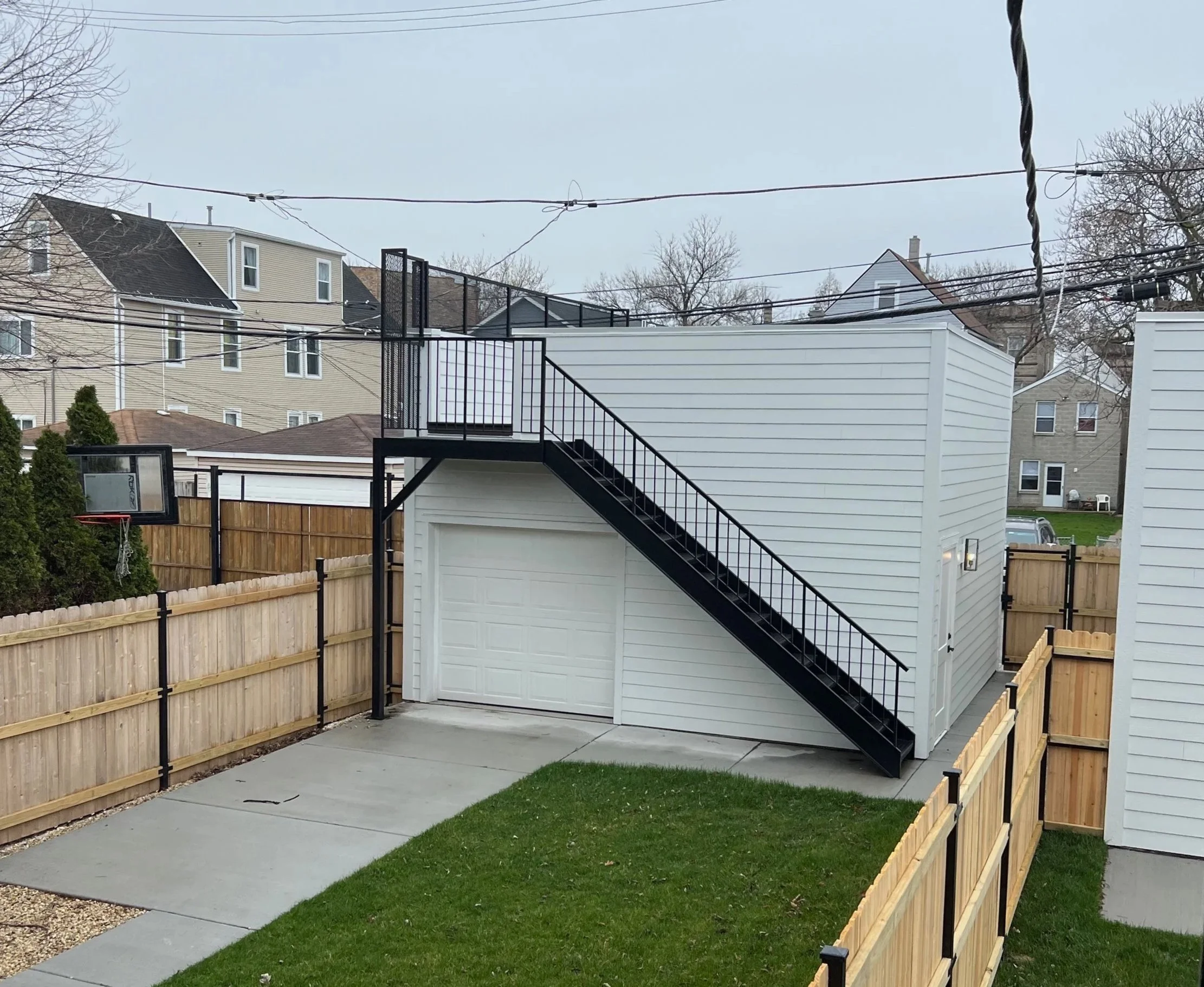Backyard with a white shed, a black staircase, a wooden fence, a basketball hoop, and neighboring houses under a cloudy sky.