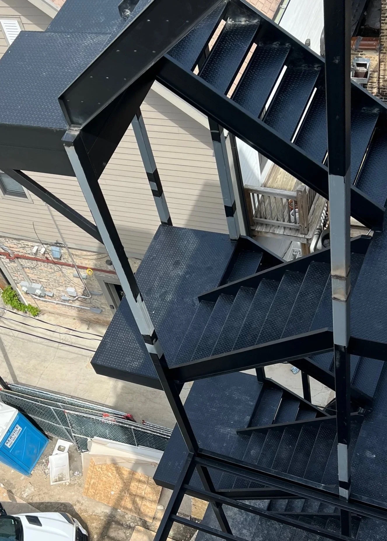The image shows a black metal fire escape staircase view from above, with outdoor residential buildings in the background, including a backyard with a wooden fence, potted plants, and a utility blue portable toilet on the street level.