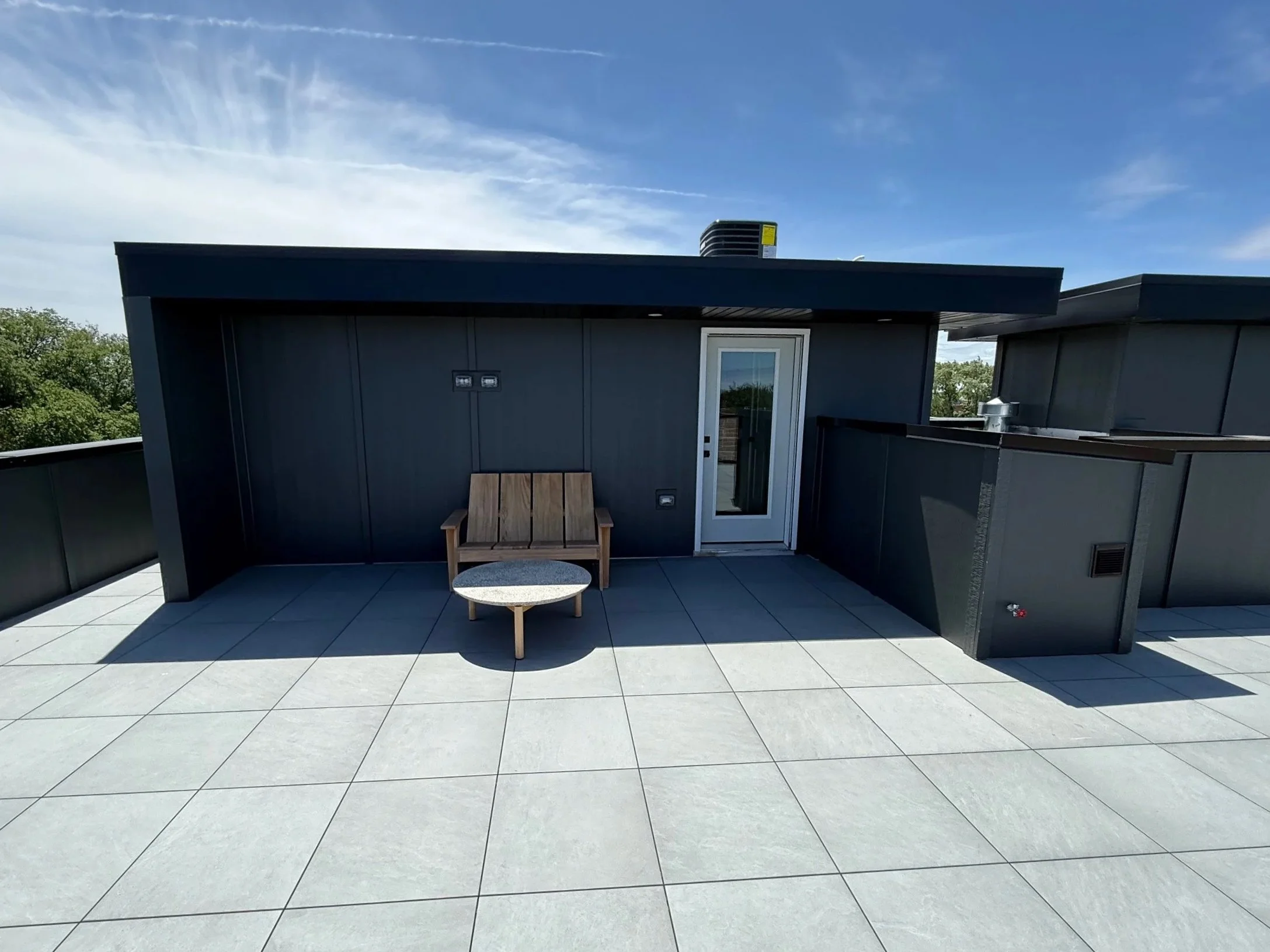 Rooftop terrace with gray tiled floor, wooden bench, small round table, and black railing, with a building exterior, blue sky, and trees in the background.
