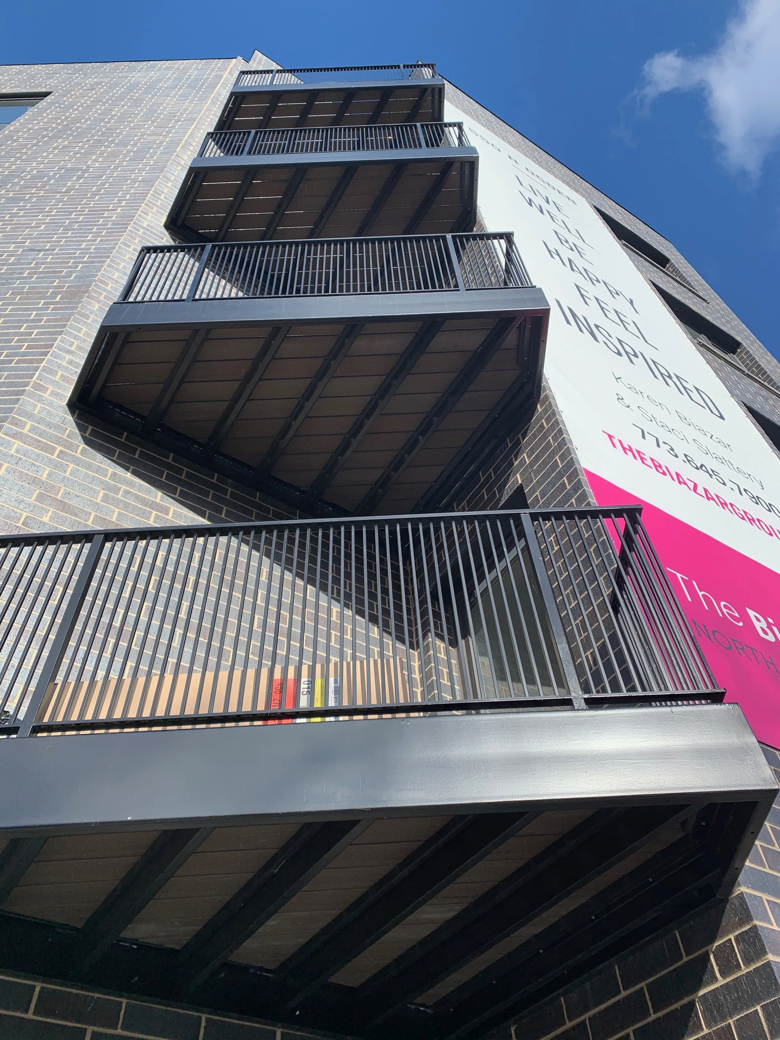Multiple black balconies on the exterior of a multi-story brick building, with a blue sky in the background and a large vertical sign on the right side of the building.