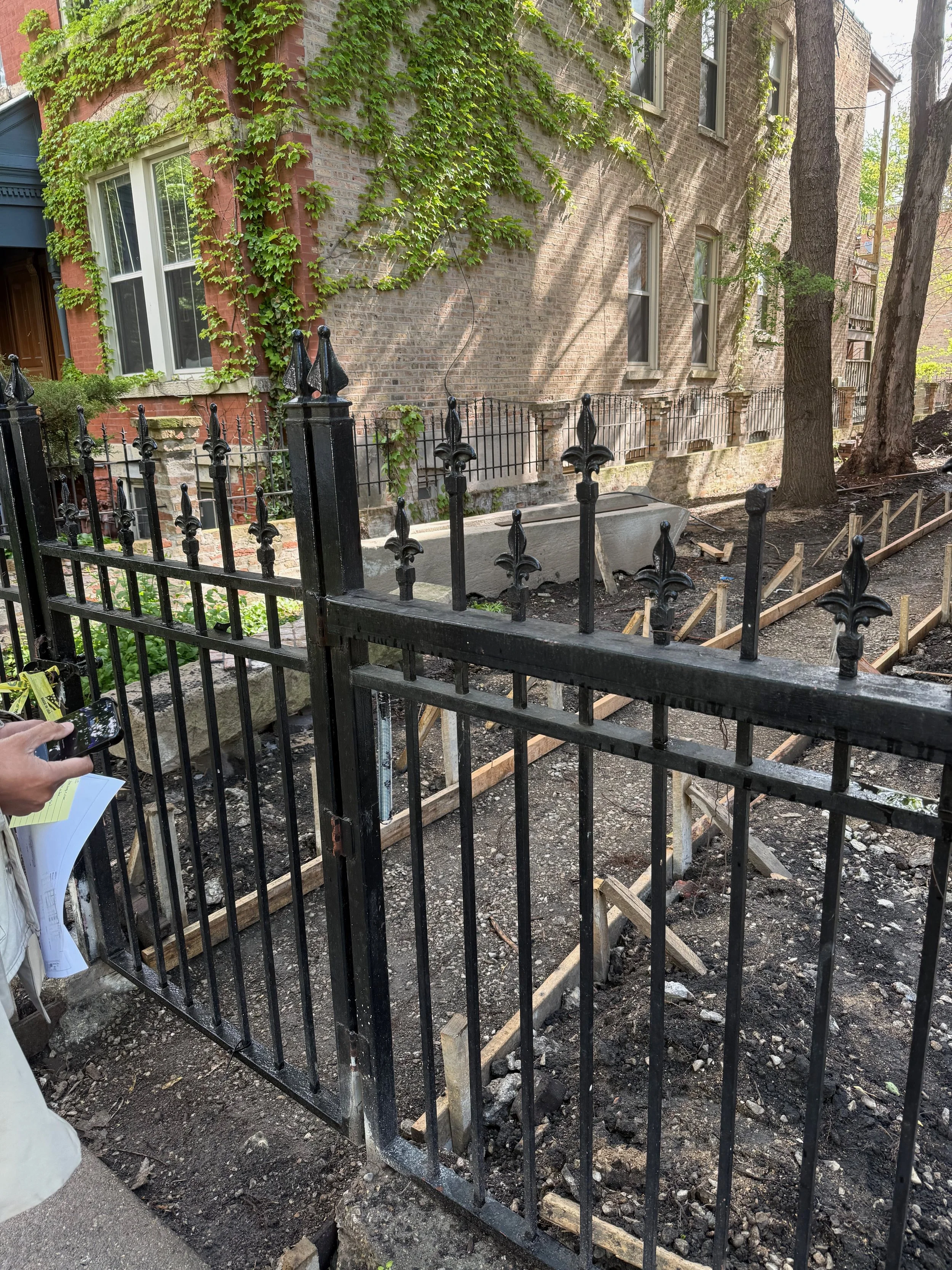 A sidewalk in front of a brick building with green ivy growing on the wall, a black iron fence, and construction work with wooden frames and dirt in the ground.