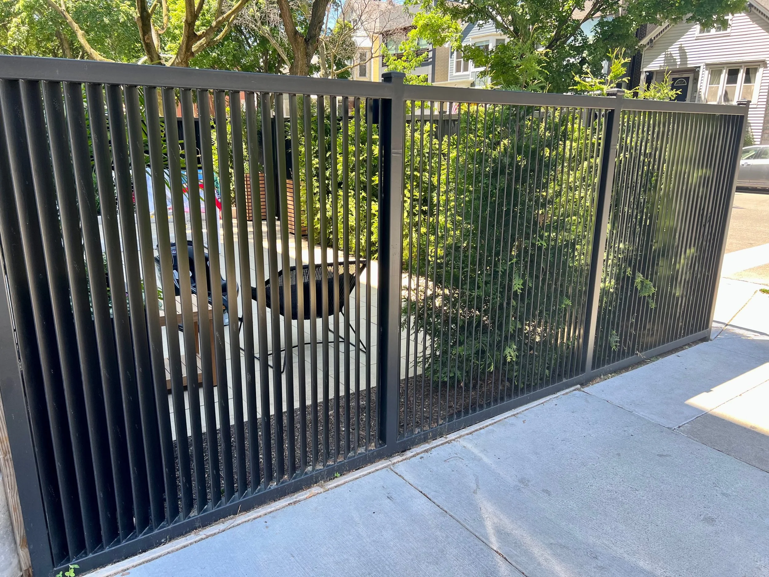 A black metal fence with vertical bars, bordering a sidewalk with concrete paving, with green trees and houses in the background.