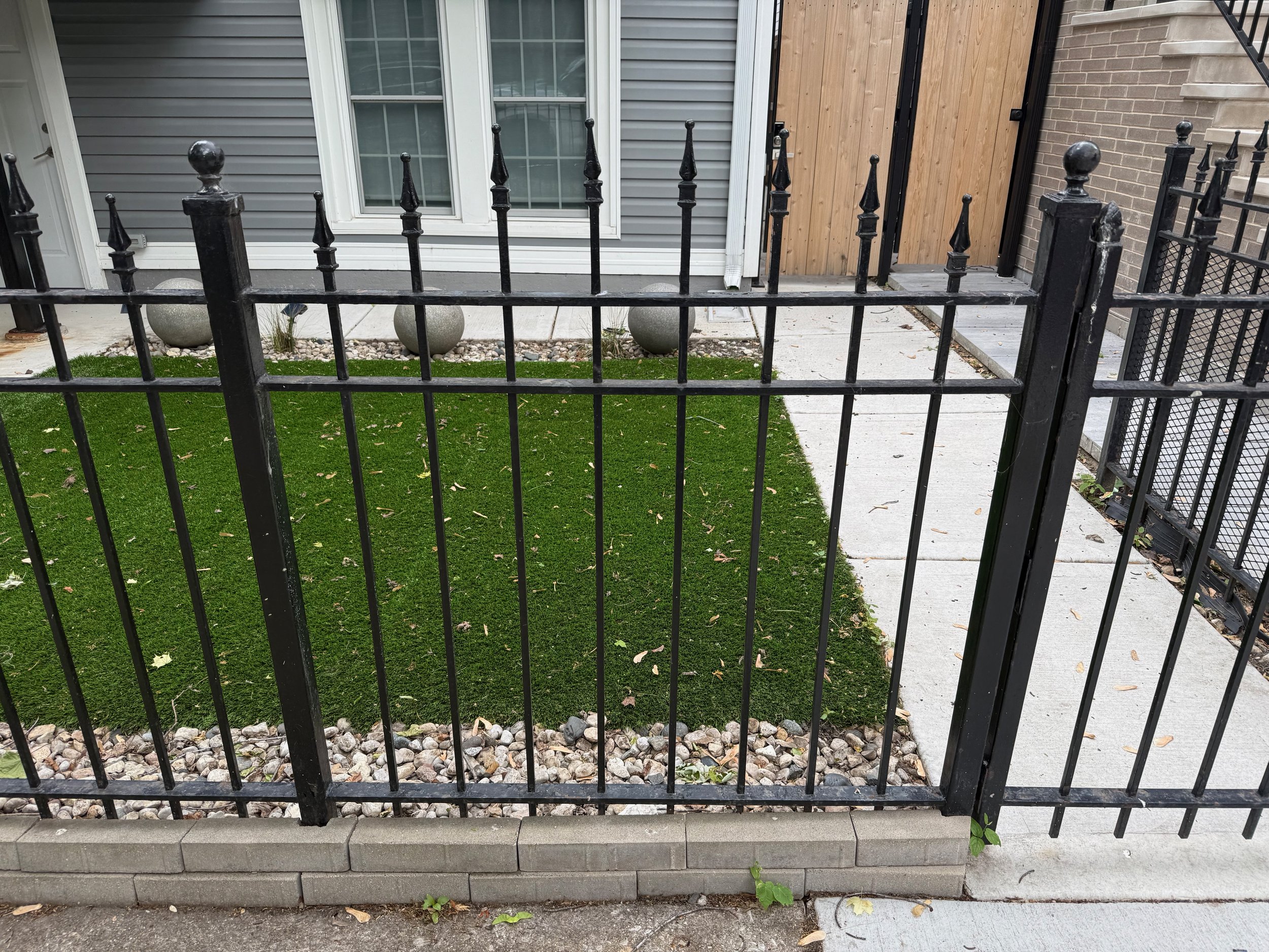 A small fenced yard with a patch of artificial grass, surrounded by a black metal fence and a narrow sidewalk. The house has gray siding, double window, and a wood panel door. There are decorative stones and spheres in the yard.
