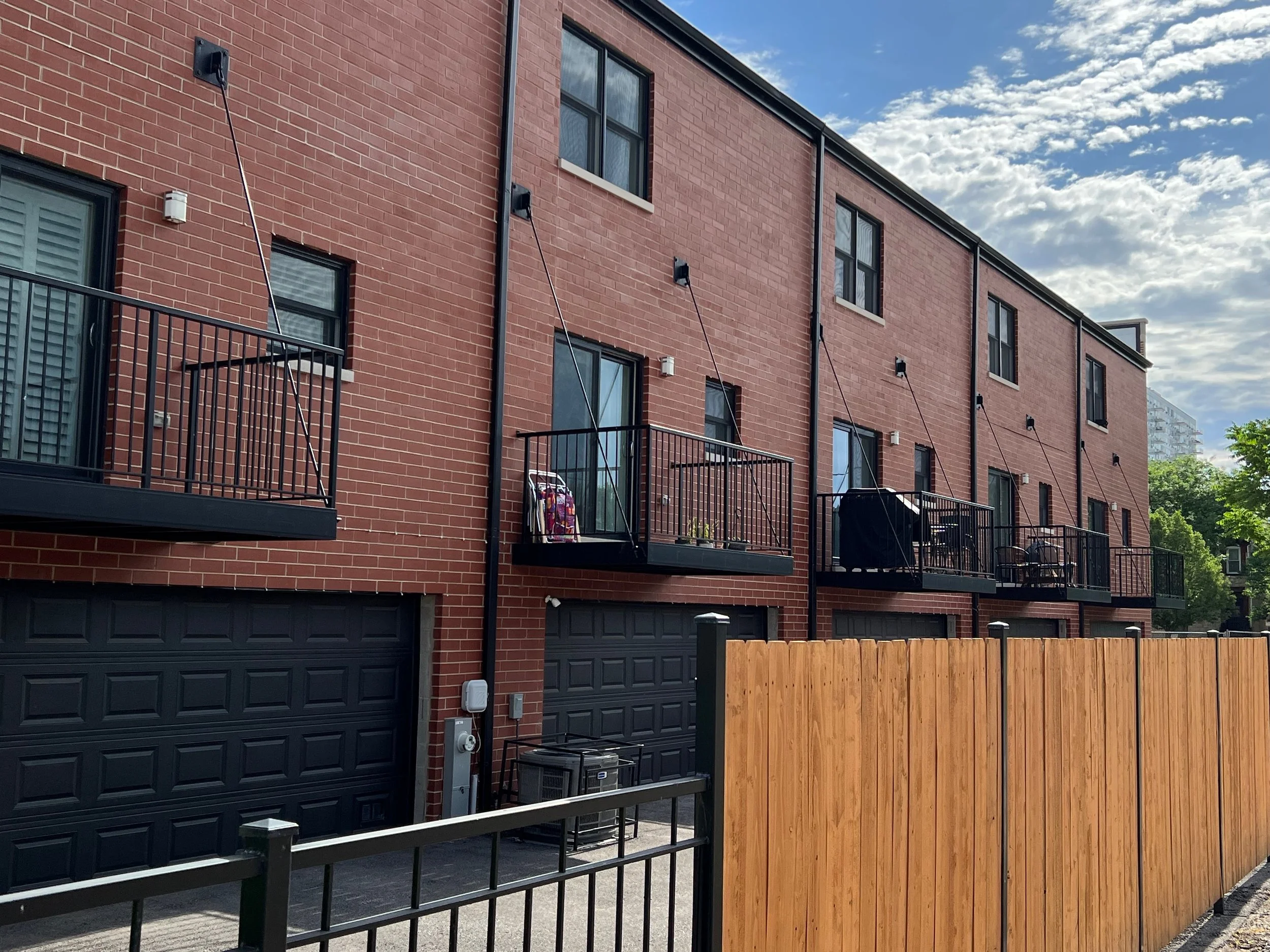 Exterior view of a modern apartment building with red brick walls, black metal balconies, and black garage doors. There are clothes hanging on one of the balconies and a wooden fence in the foreground. The sky is partly cloudy with sunlight illuminat