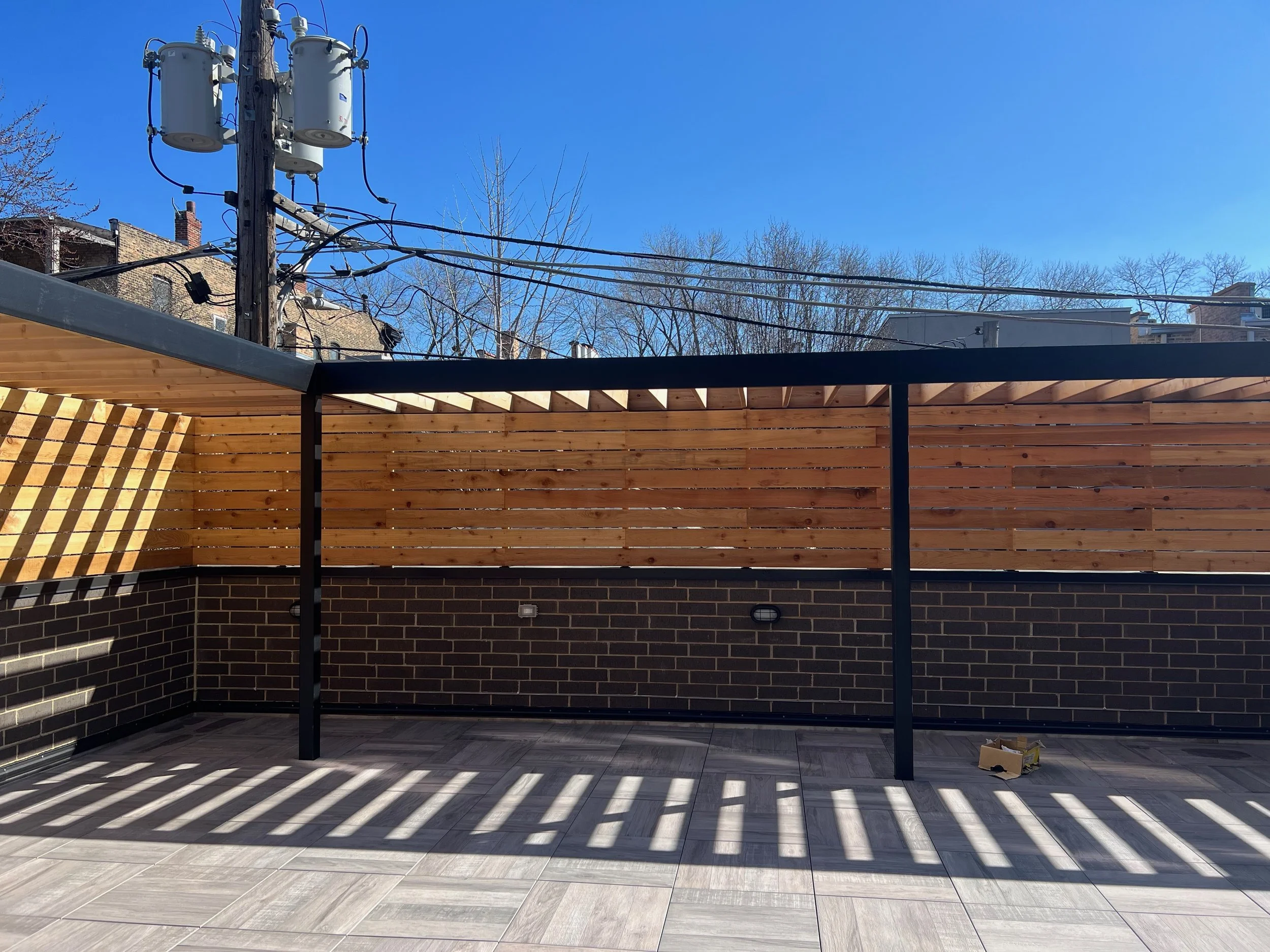 An outdoor space under construction with a wooden fence, brick wall, and wooden floor. Shadows of a railing cast striped patterns on the floor. Electrical poles and wires are visible against a clear blue sky.