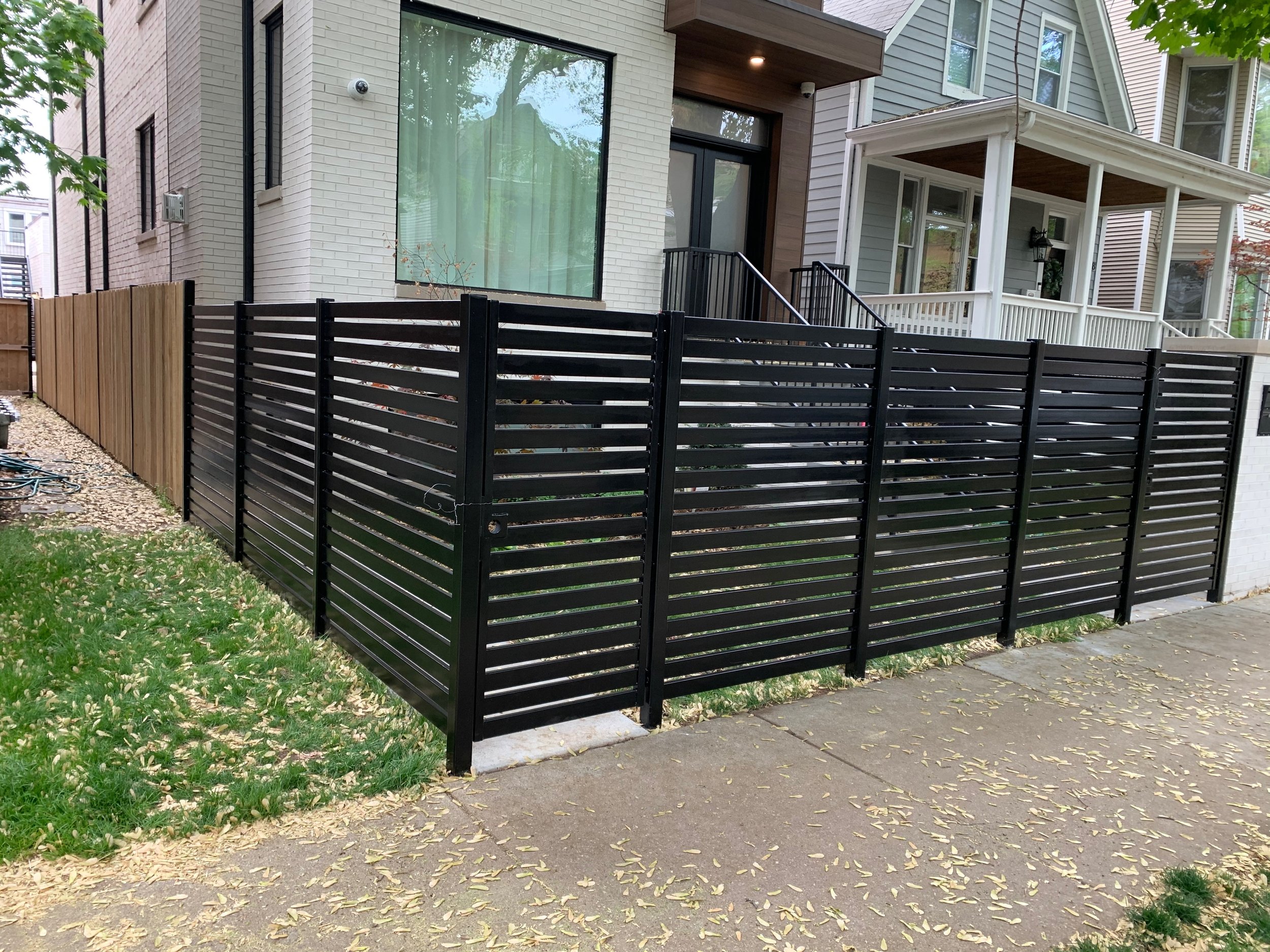 A modern house exterior with a large window, a porch, and a black slatted fence. There is a lawn with fallen yellow leaves, a sidewalk, and trees around.