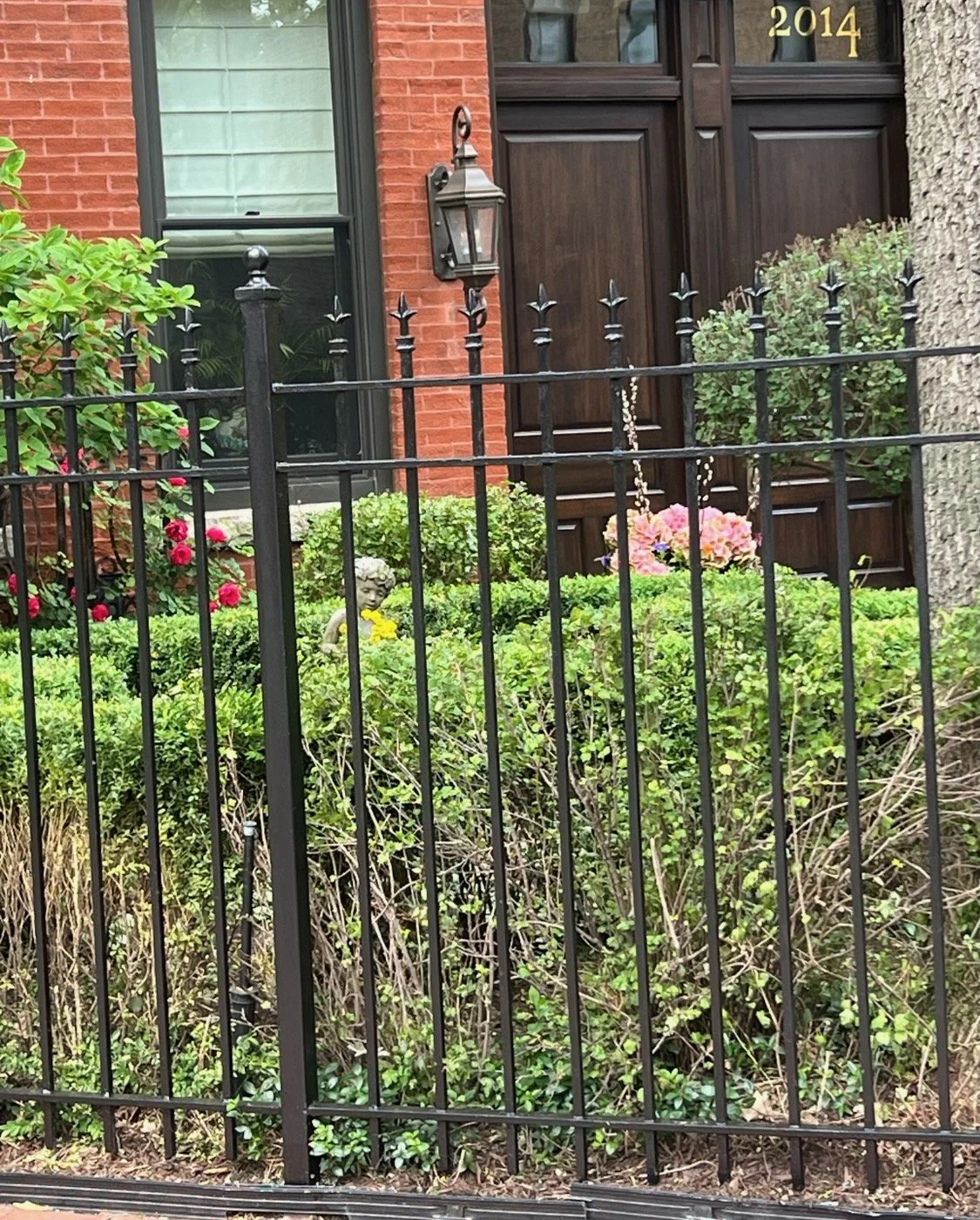 Front yard of a brick house with a black metal fence, green bushes, pink flowers, a small statue of a child, and a dark wooden door with the number 2014.
