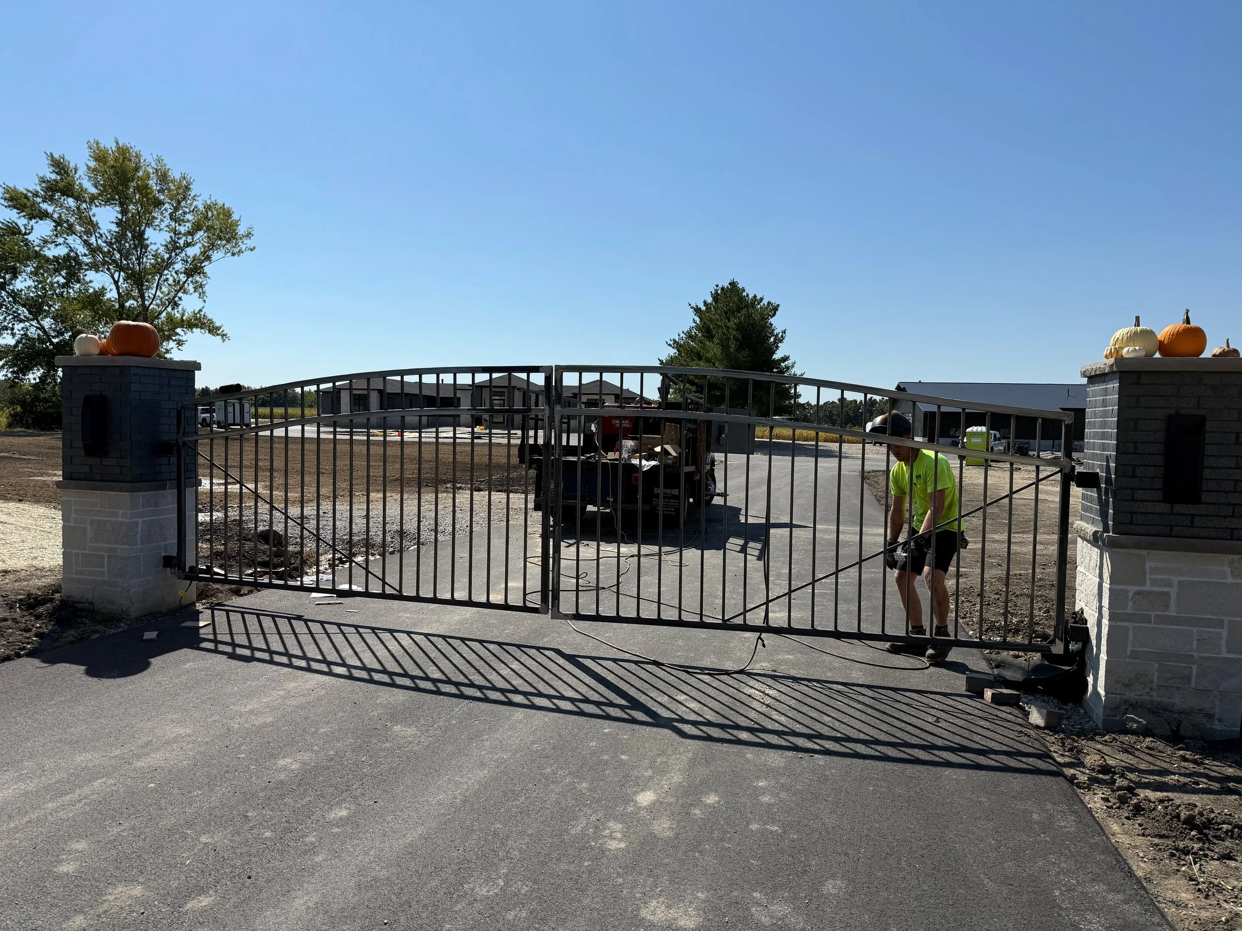 A construction worker in a neon yellow shirt working near a metal gate at a construction site, with concrete pillars topped with pumpkins on either side and trees in the background under a clear blue sky.