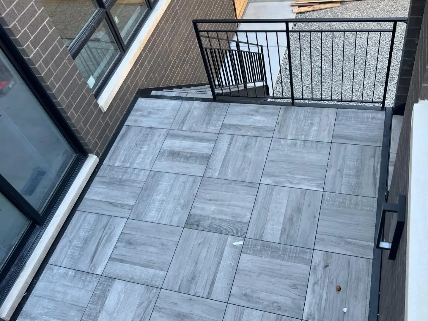 Small balcony with gray tile flooring, black railing, brick walls, glass windows, and a white ledge.