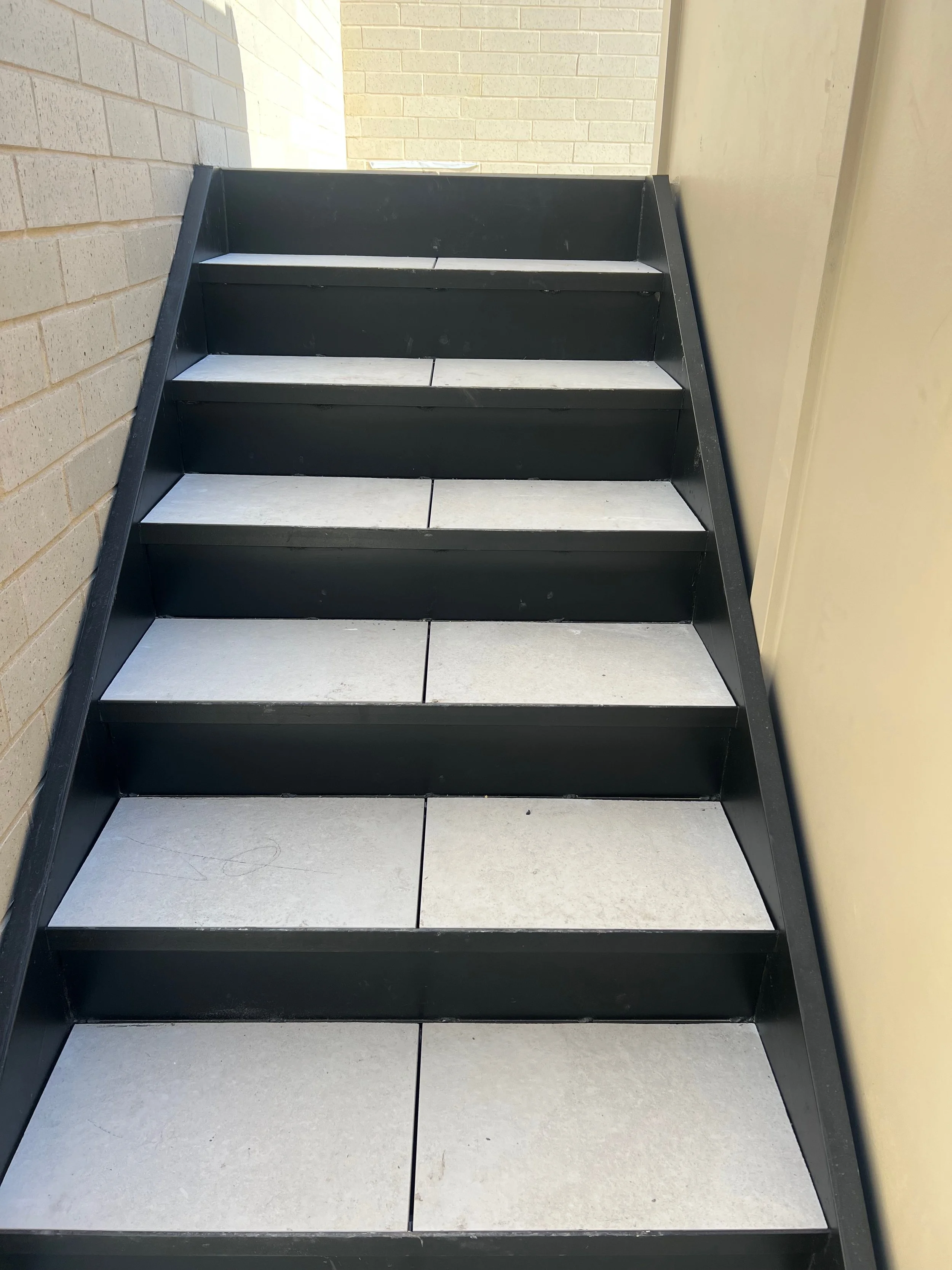 Black metal stairs with white tile steps against beige brick wall and yellow wall in the background.