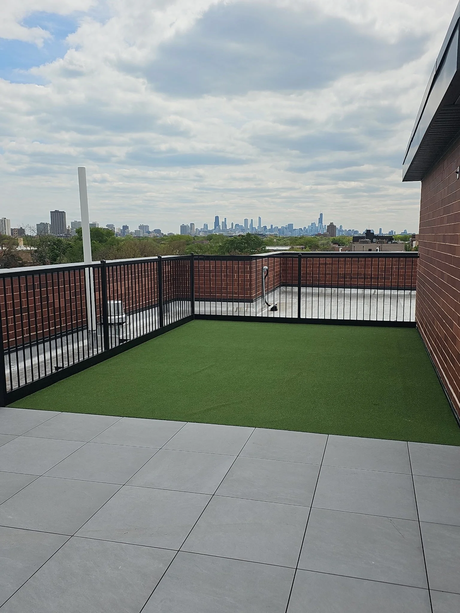 Image of a rooftop patio with gray tile flooring, a green artificial grass section, a black metal railing, and a city skyline in the background under a partly cloudy sky.