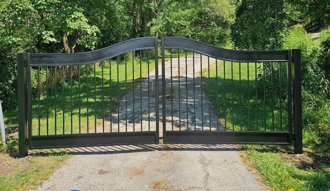 Black metal gate with curved top, closing a gravel driveway surrounded by green trees and grass.